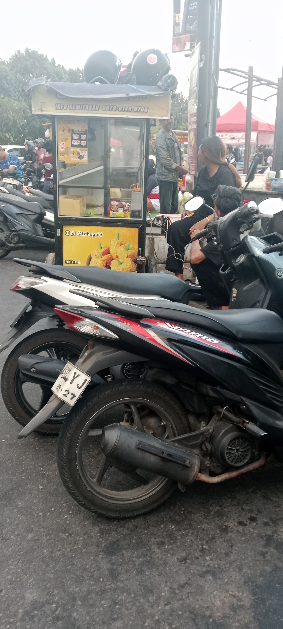 In Ngemplak, Indonesia, on an overcast day, a street food cart selling fried food is positioned at the side of a road, with two helmets resting on top of the cart. The cart features a menu with prices for various fried food items, along with contact information for potential business partners. A woman in a black shirt is visible in the background, looking away from the camera, while a man wearing a denim jacket stands near the cart. Several motorcycles are parked in front of and around the cart, with one prominently displayed in the foreground, its license plate showing "AG 4632 YJ 09.27". The overall scene depicts a typical street scene with food vendors and customers.
