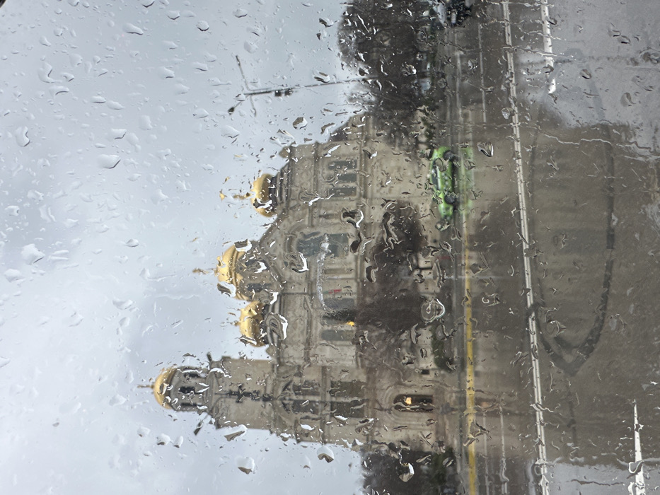 The image depicts a rainy day scene in Varna, Bulgaria, viewed through a window covered in numerous water droplets. The central subject is the Dormition of the Theotokos Cathedral (Cathedral of the Assumption of the Virgin), a large, light-colored stone structure with multiple ornate golden domes and a prominent bell tower, positioned beyond a multi-lane wet asphalt road. The sky above is uniformly overcast and grey. On the wet road in the midground, a distinct lime green vehicle, possibly a taxi with visible markings, is parked or moving. Reflected light from the wet road surface indicates a high moisture level. Bare deciduous trees or shrubs line the area between the road and the cathedral. A street light pole is visible on the far right. No discernible human figures are clearly visible.