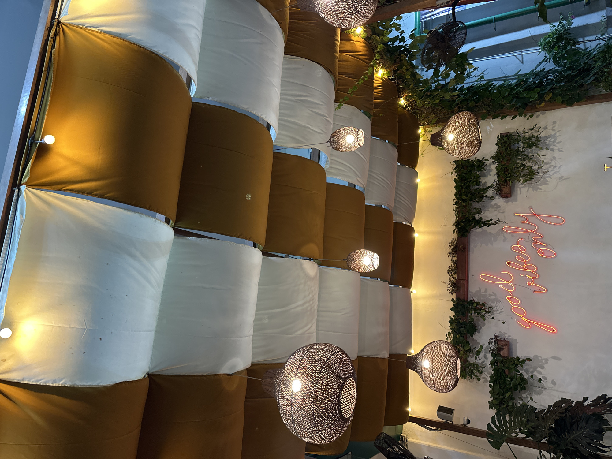 This media file captures an interior scene, likely a cafe or restaurant, with a distinctive decorative wall. The wall is constructed from alternating cylindrical elements covered in brown and white fabric, creating a textured, undulating surface. Interspersed throughout the scene are several hanging light fixtures. Some are globe-shaped with intricate woven shades, emitting a warm glow, while others are simple, exposed bulbs. Green vines and foliage are strategically placed, draped over the wall and near the ceiling, adding a natural element to the space. A ceiling fan is visible in the upper portion of the frame.
On the right side of the image, a white wall serves as a backdrop for a neon sign. The sign, in a vibrant coral or pink hue, spells out the phrase "good vibes only." The lighting suggests it is evening or the interior lighting is intentionally dimmed, creating a cozy and ambient atmosphere. The overall impression is one of a trendy, stylish, and inviting establishment. The location context of Kuta Selatan, Indonesia, suggests a tropical or subtropical climate, which might influence the choice of lush greenery in the decor. No people are visible in the frame.