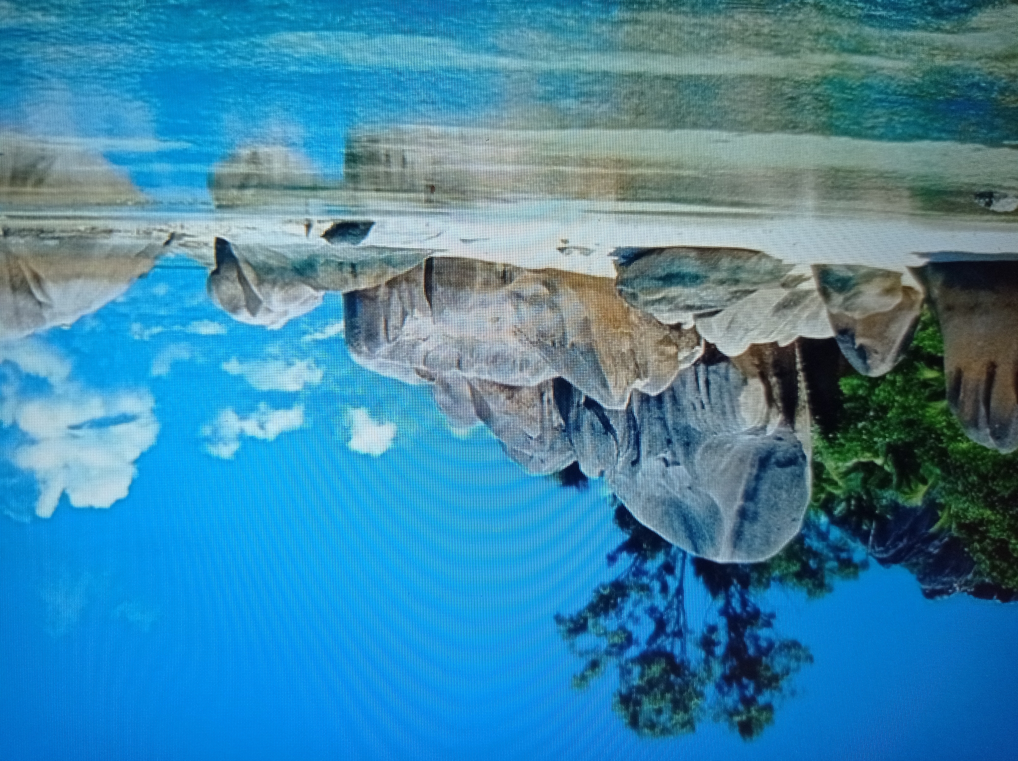 This is a photograph of a tropical beach scene. The foreground is dominated by clear, shallow water reflecting the sky and the shoreline. The shoreline itself is sandy and scattered with large, smooth, grey and beige boulders. In the background, more boulders are visible, some covered in lush green vegetation and trees. The sky is a vibrant blue with a few fluffy white clouds. There is no indication of the city of Mukachevo, Ukraine, in this image; it depicts a natural beach environment.