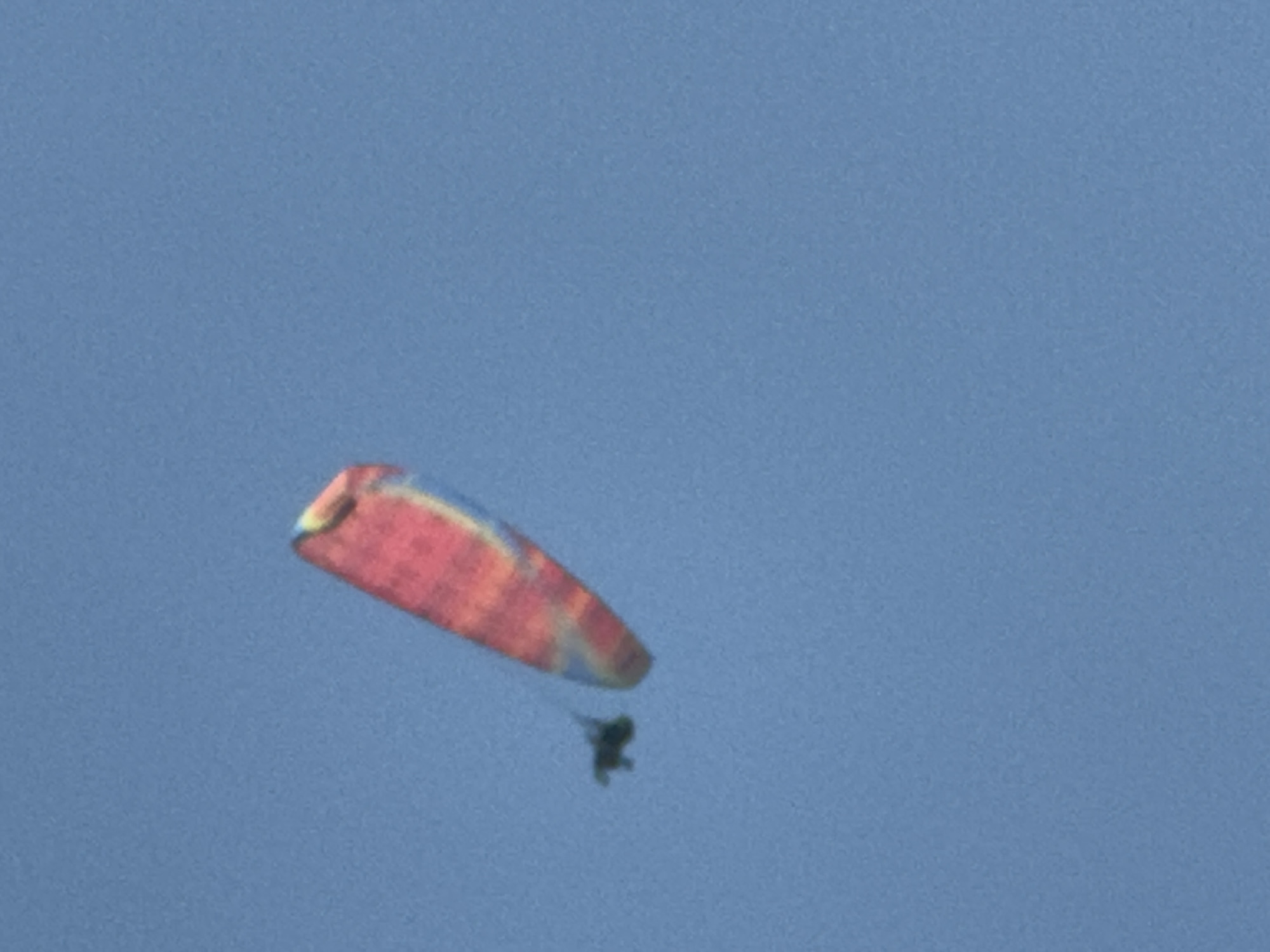 A paraglider with a colorful red, yellow, and blue wing is visible against a clear blue sky. The paraglider appears to be in motion, with the wing extended and the pilot suspended beneath it. The image is taken from a low angle, looking up at the paraglider. The location is not specified in the city of Boreti, Montenegro, as there is no visual information to support this.