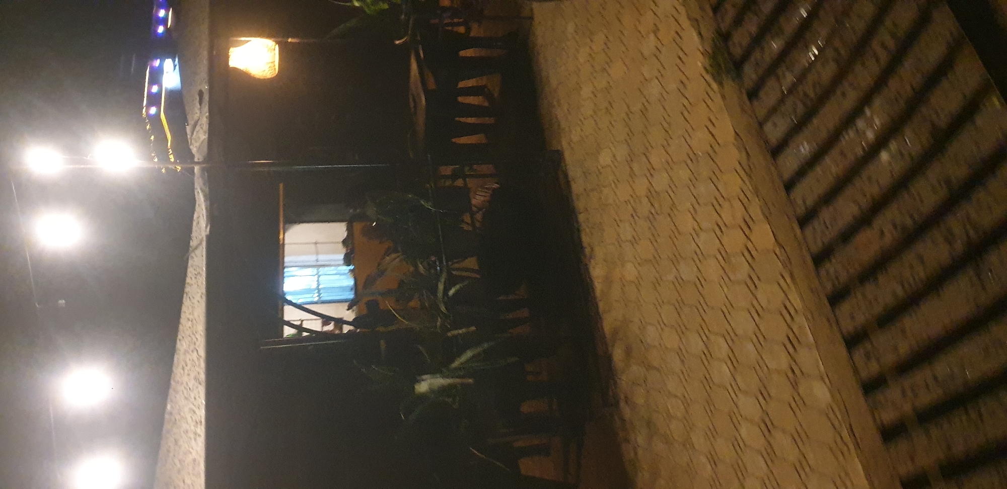 The image captures an outdoor seating area at night, likely a restaurant or bar, illuminated by string lights and a hanging lamp. The ground is paved with hexagonal tiles. In the background, there is a building with a window covered by light blue curtains. Several dark, shadowy figures of people are visible sitting at tables. There are also plants with large leaves in the foreground. The overall impression is that of a dimly lit, casual outdoor gathering space. The location is identified as Narayi, Nigeria.