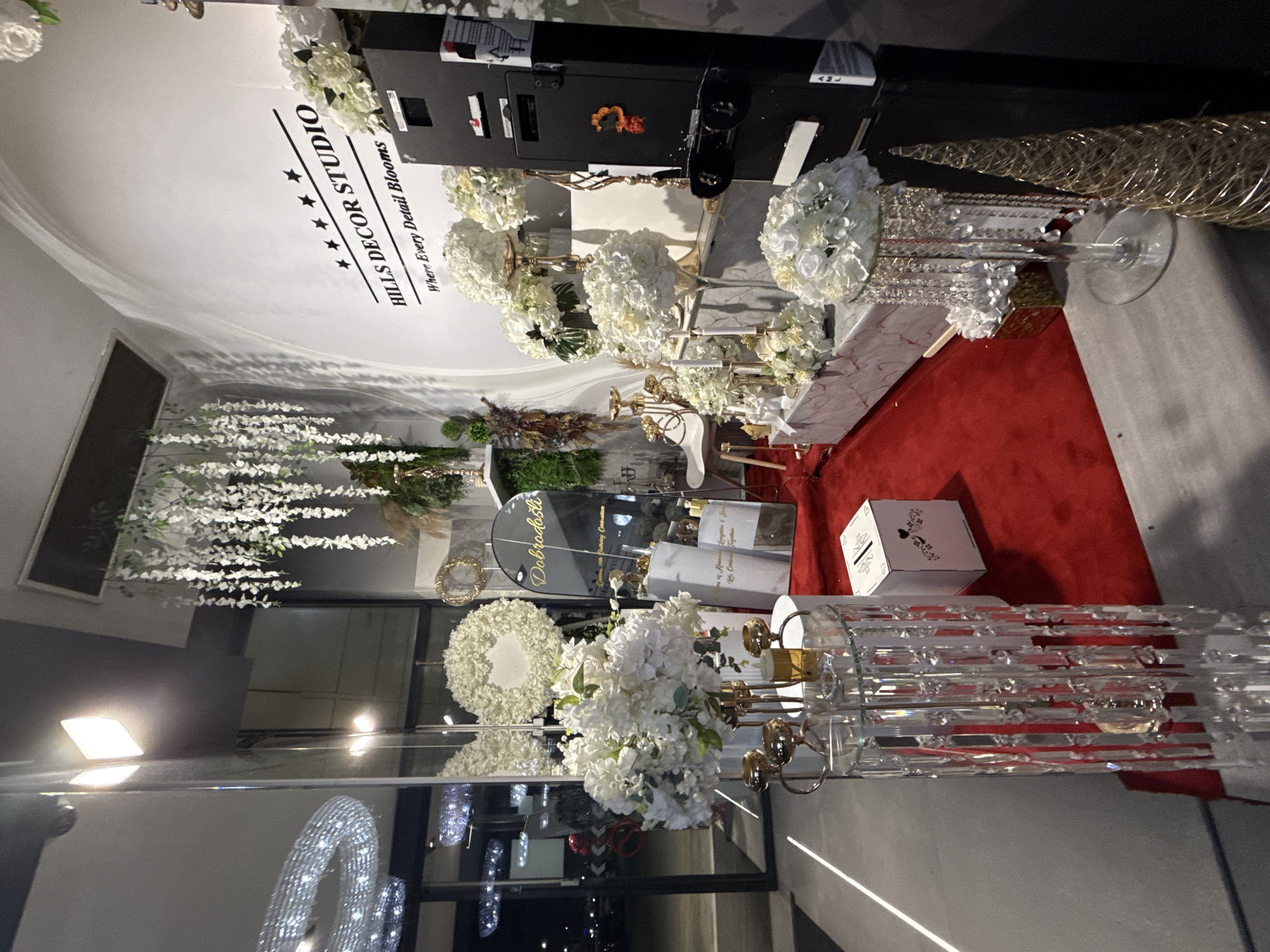 The image depicts the interior of a store, possibly a wedding or event decor studio, with a prominent sign on the wall reading "HILLS DECOR STUDIO Where Every Detail Blooms." The store is filled with white floral arrangements, crystal chandeliers, and decorative items, set against a white wall and a red carpet. In the foreground, a clear glass stand with hanging crystals holds a bouquet of white flowers. To the right, a tall black cabinet with several compartments is visible, and next to it is a golden, cone-shaped decorative object. The store appears to be located in Ilidža, Bosnia and Herzegovina, as suggested by a reflective surface displaying the word "Dobrodošli" (Welcome in Bosnian) and text referencing "Goran's 50th Birthday Celebration." No people are present in the image.