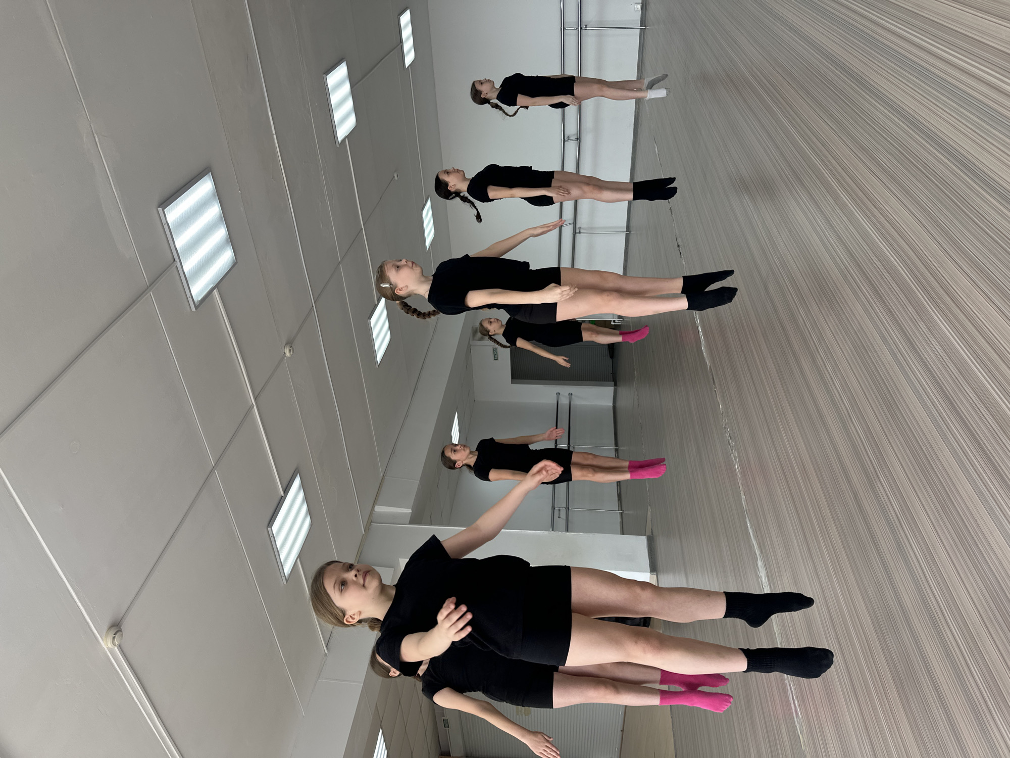 A group of six young girls are in a dance studio in Votkinsk, Russia. The girls are dressed in black leotards or t-shirts and shorts, and they appear to be practicing a dance routine. Some are wearing socks in colors of black, pink and grey. The studio has a mirrored wall along one side, where the girls are lined up, allowing them to see their reflections. The floor is a light wood with linear grain. Above, fluorescent lights are inset in a white tiled ceiling.