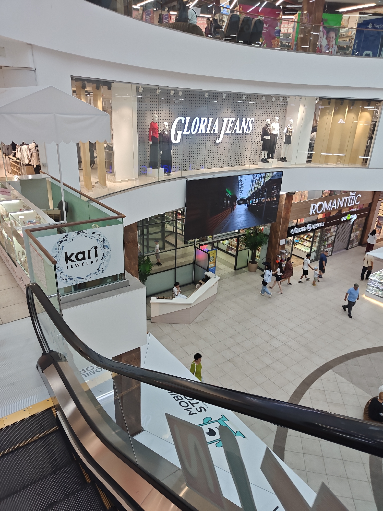 The image shows an interior view of a multi-level shopping mall, likely in Petropavl, Kazakhstan. An escalator with black handrails and metal steps dominates the foreground, leading upwards. The mall features a bright, modern aesthetic with white walls and polished tile flooring.
Several storefronts are visible. On the left, a "Kari Jewelry" store displays its logo on a glass partition. Further back, the "Gloria Jeans" clothing store has a large sign and mannequins in its window. Across the walkway, the "Romantic" flower shop and other businesses are open.
People are seen walking through the mall. A woman in a green top walks near the escalator. A family, including two adults and a young child, are walking towards the "Romantic" store. Several other shoppers are scattered throughout the concourse. The mall appears to be moderately busy. Natural light streams in from upper levels, and artificial lighting illuminates the interior. Promotional displays and signage are visible throughout the space.