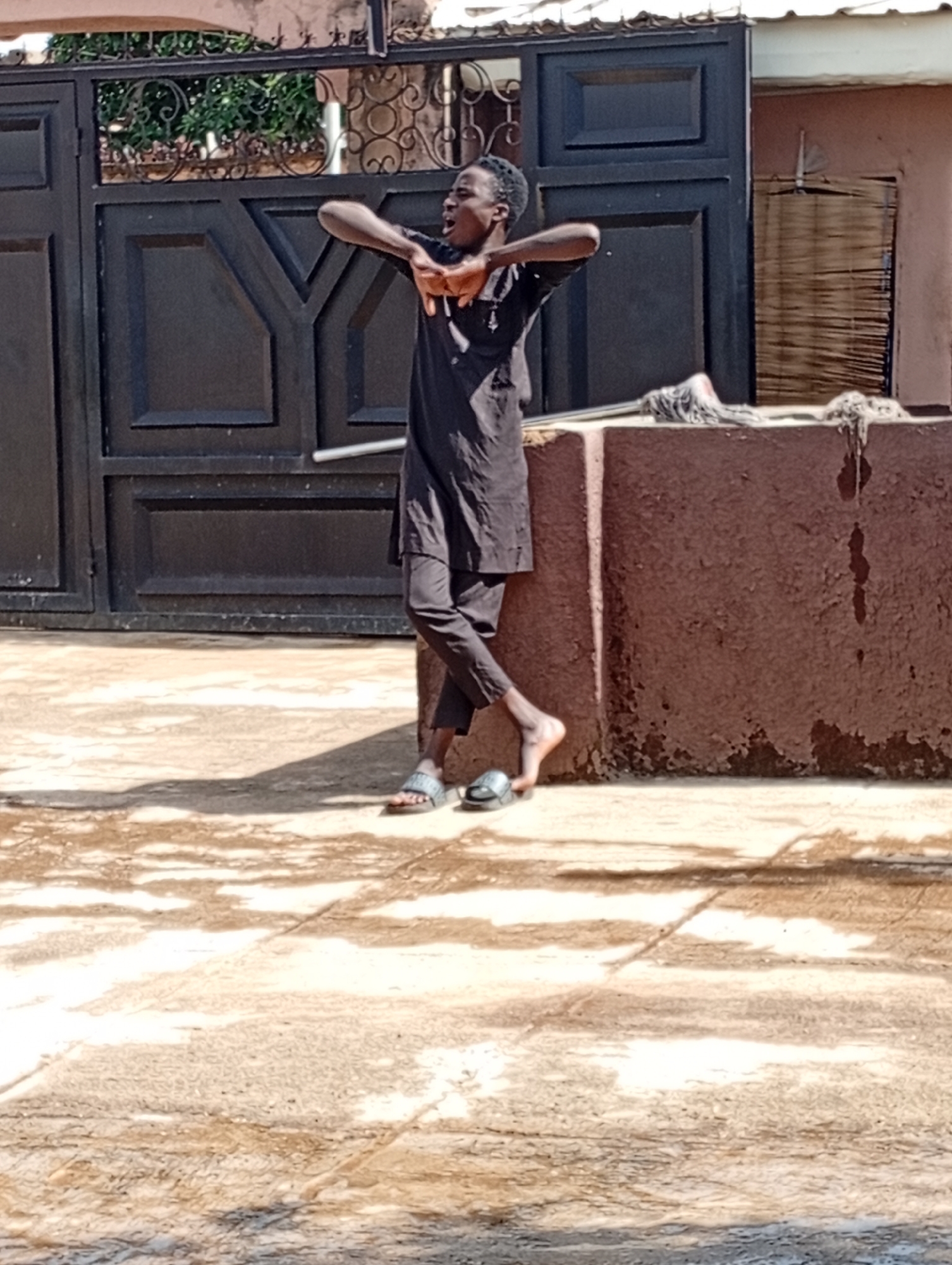 The image captures a young Black male standing in front of a dark metal gate in Jos, Nigeria. He is wearing a dark, loose-fitting outfit, possibly traditional attire, with pants that are tucked into his sandals. His arms are raised and bent at the elbows, with his hands positioned near his head, and his mouth is open as if speaking or exclaiming. His right leg is crossed over his left, and he is wearing simple slide sandals. The ground beneath him appears to be paved and wet, with sunlight creating distinct shadows. To his right, a reddish-brown, rough-textured wall or structure is visible, topped with a few items that could be cleaning supplies. The gate is large and ornate, suggesting it might be an entrance to a residential or commercial property. The overall scene depicts a moment of energetic expression in an outdoor urban setting.