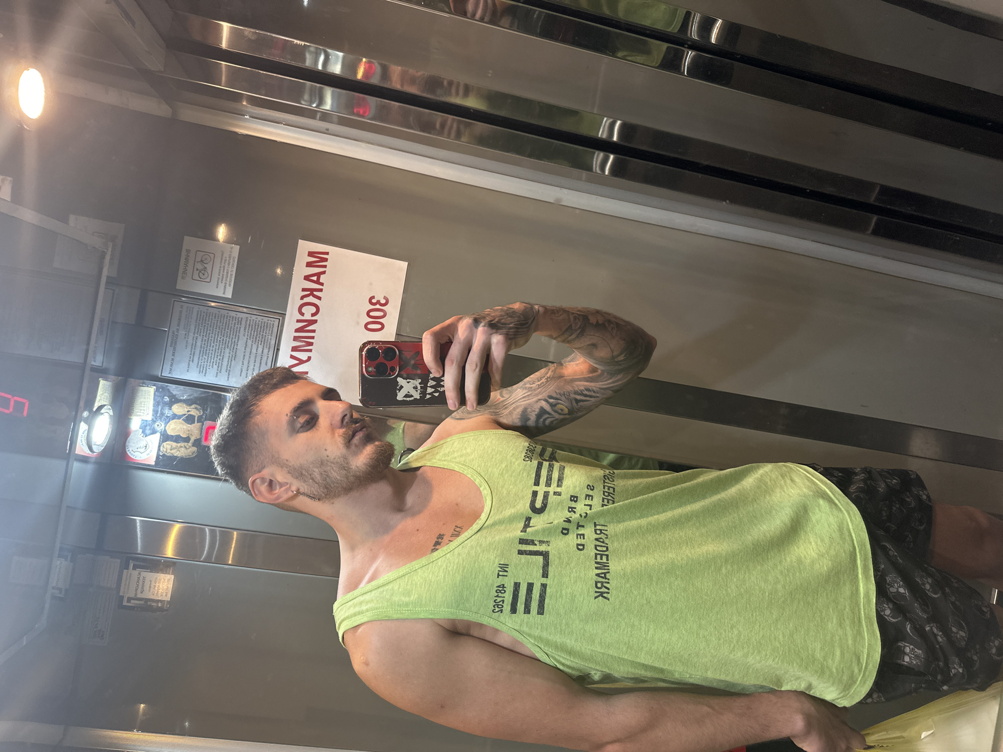 A man takes a selfie in an elevator. The elevator has metal walls and a sign that reads "MAKCИMAЛHO 300" which translates to "MAXIMUM 300". The man is wearing a green tank top and shorts. His left arm is tattooed. The elevator is likely in Sofia, Bulgaria due to the Cyrillic alphabet on the sign.
