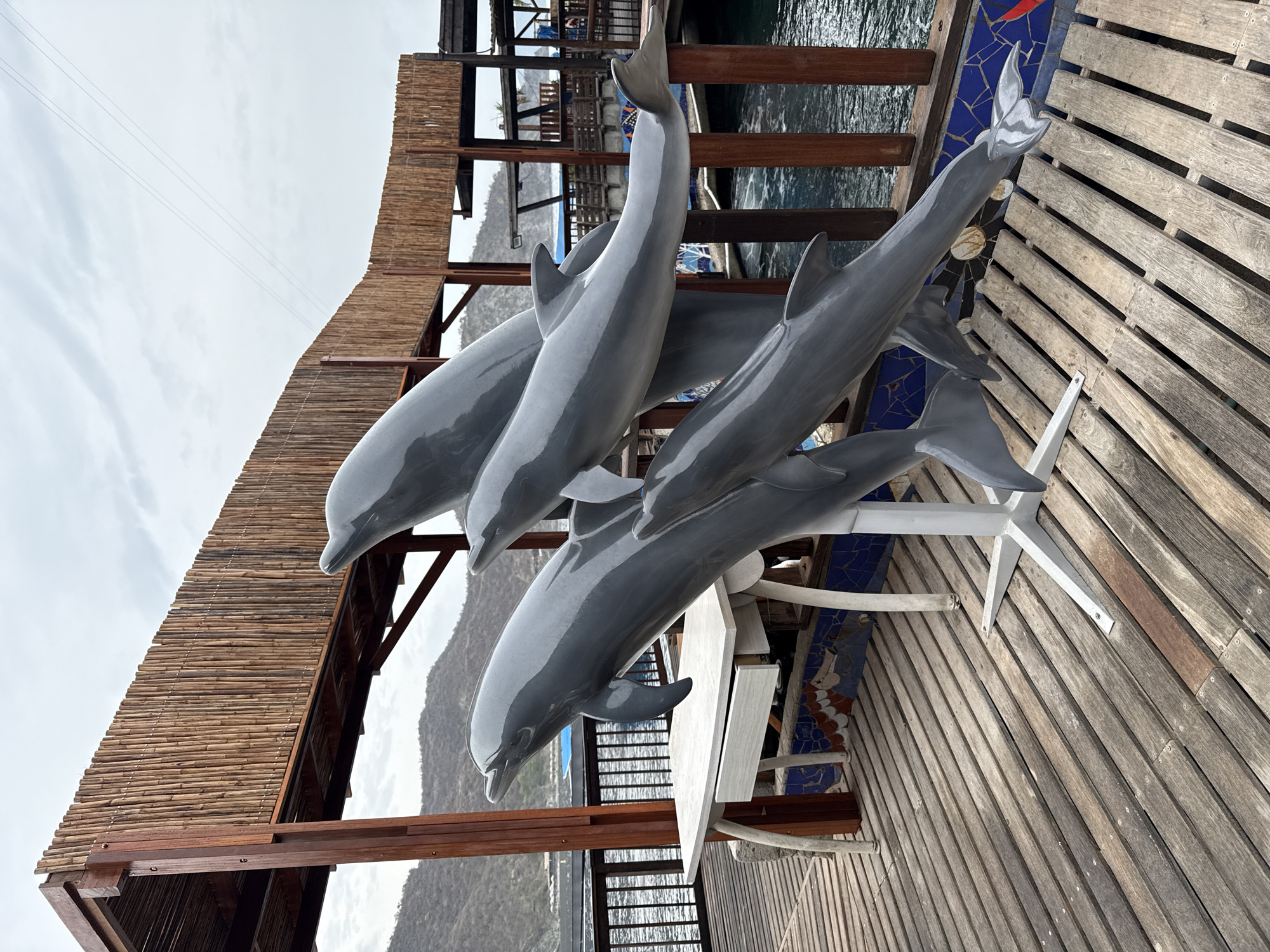 A gray dolphin sculpture, comprised of four dolphins in a leaping formation, stands on a wooden pier in Santa Marta, Colombia. The sculpture is mounted on a white metal base. In the background, a building with a bamboo-covered roof and wooden supports is visible. Beyond the building, a body of water with a distant hilly landscape under a cloudy sky can be seen. A white table with a drawer is positioned to the left of the sculpture. The pier is made of weathered wooden planks.