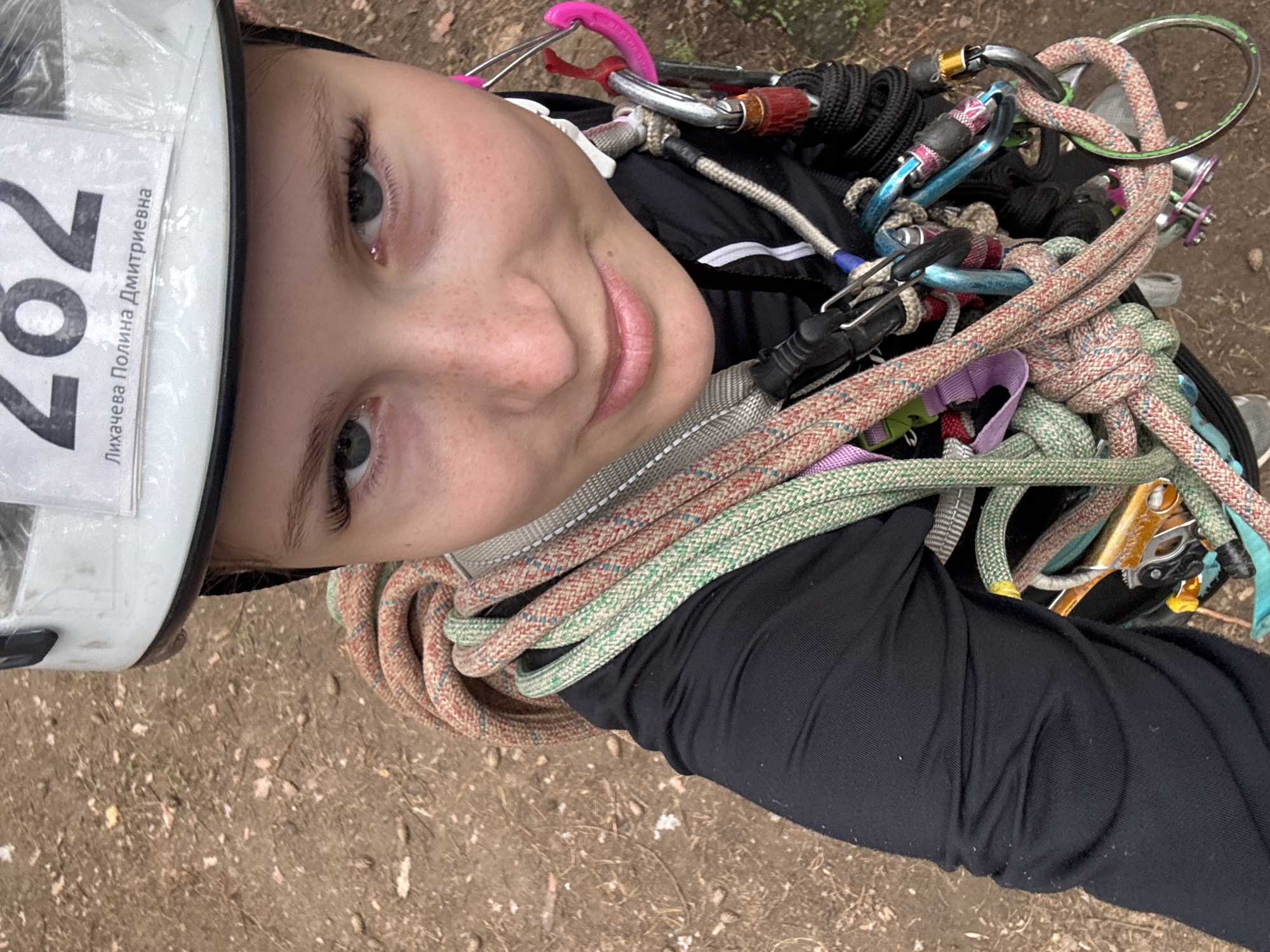 This is a close-up, vertically oriented, outdoor photograph of a young girl wearing a white helmet. The girl has light skin with freckles and is looking directly at the camera with a slight smile. Her eyes are open and her eyelashes are prominent. She is wearing a black long-sleeved shirt.
Around her neck and chest, the girl is wearing a harness and is surrounded by climbing ropes and carabiners in various colors including red, white, green, blue, and pink. Some of the carabiners appear to be metal, while others have colored plastic elements. The ropes are coiled and some are knotted.
The setting appears to be outdoors on a dirt ground with some dry leaves and twigs. The lighting suggests it is daytime, likely sunny, as there are shadows and the overall image is well-lit.
Visible text on the helmet reads "702" and "Лихачева Полина Дмитриевна". This indicates the girl's name is Polina Dmitrievna Likhacheva and she has the number 702.
The overall impression is of a participant in an outdoor climbing or adventure activity, possibly a competition or training event, given the bib number and equipment. The girl appears confident and engaged. The location context is Chaikovsky, Russia.