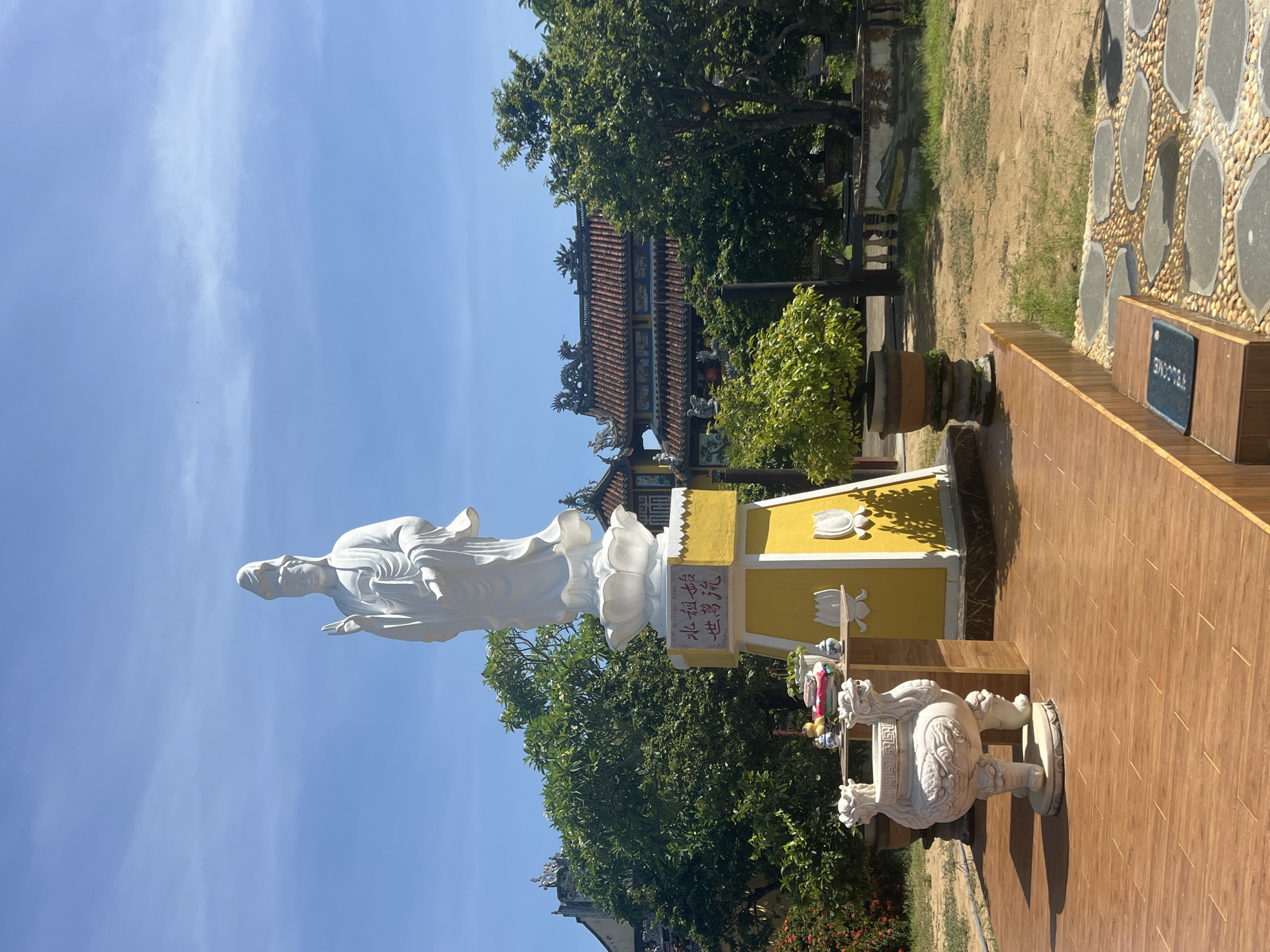This is a vertical, eye-level, full shot of a white statue of a person standing on a lotus flower, with a yellow pedestal behind it. The statue is adorned with Chinese writing on the pedestal. To the right of the statue is a large white sculpture of a mythical creature that resembles a dragon or lion. In the background, there is a traditional Vietnamese building with a tiled roof and intricate decorations, surrounded by lush green trees. The sky above is clear blue with some wispy clouds. The scene appears to be in a temple or garden setting in Đà Nẵng, Vietnam, given the architectural style and the presence of Buddhist imagery. The lighting suggests it is daytime, likely sunny. The overall mood is serene and contemplative.