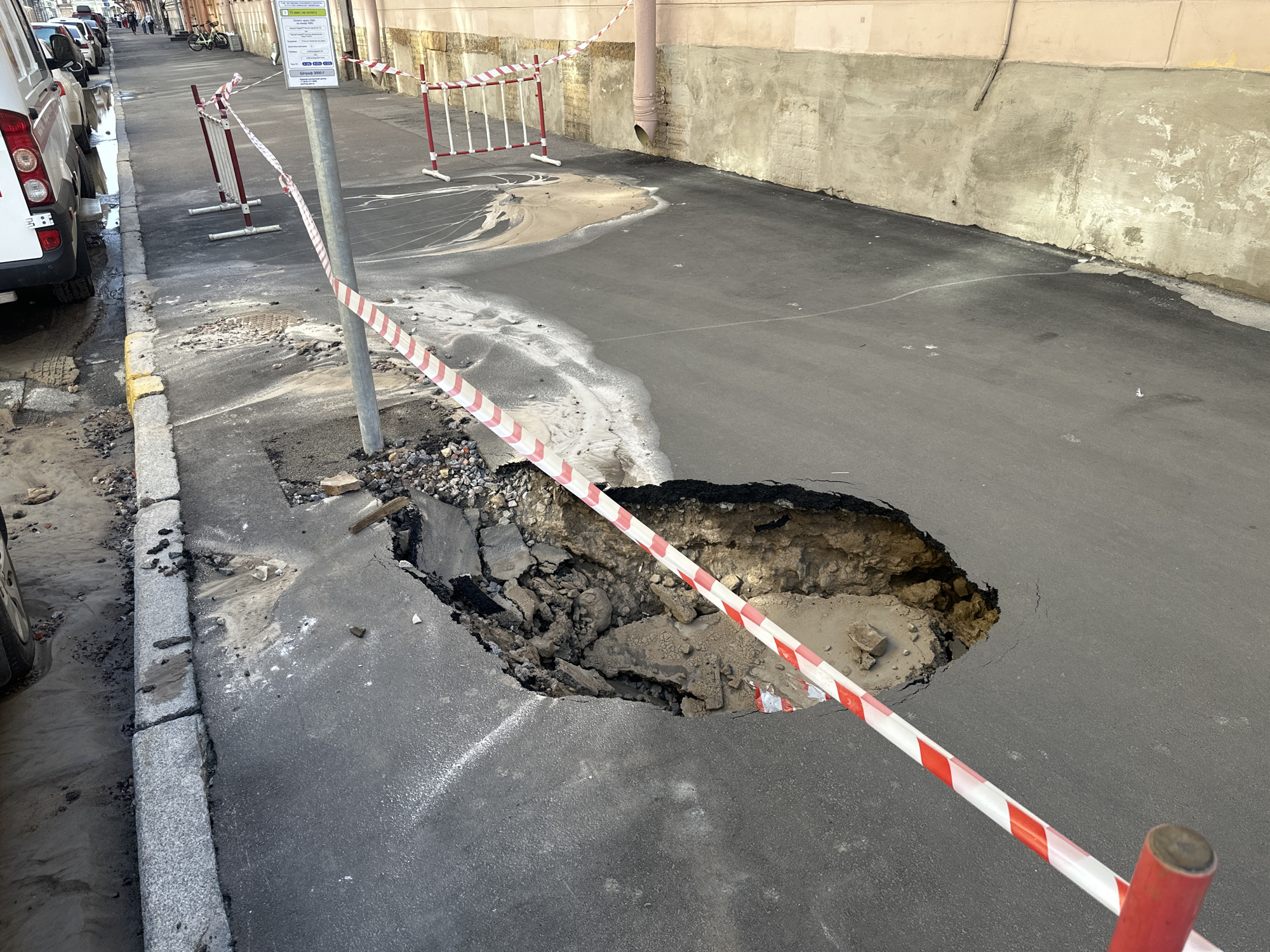 The image shows a large sinkhole in an asphalt street in Saint Petersburg, Russia. The sinkhole is bordered by red and white striped caution tape and a metal barrier. The hole exposes broken asphalt and debris, with a pipe visible at the bottom. A white van is parked on the left side of the street. Other vehicles and pedestrians are visible in the background. The street is lined by buildings with weathered facades.