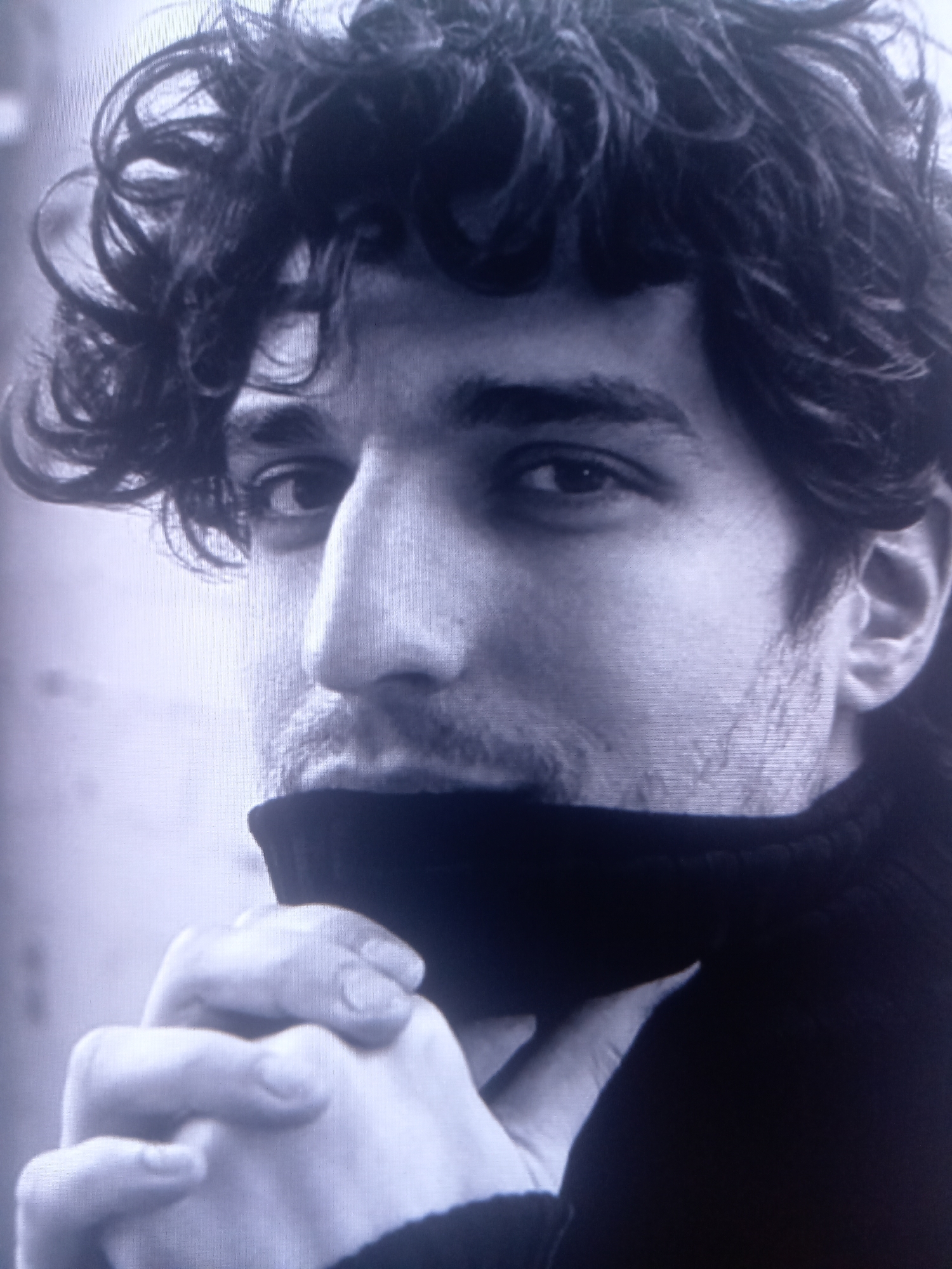 A black and white, close-up, waist-up shot of a young adult male, seen from a slightly low angle. The male has dark, curly hair, prominent eyebrows, and a slight stubble. He is wearing a dark turtleneck sweater, and his hands are clasped in front of his neck, partially obscured by the sweater. The background is bright and out of focus, suggesting an outdoor setting. The lighting is soft, with the subject's face illuminated from the front.