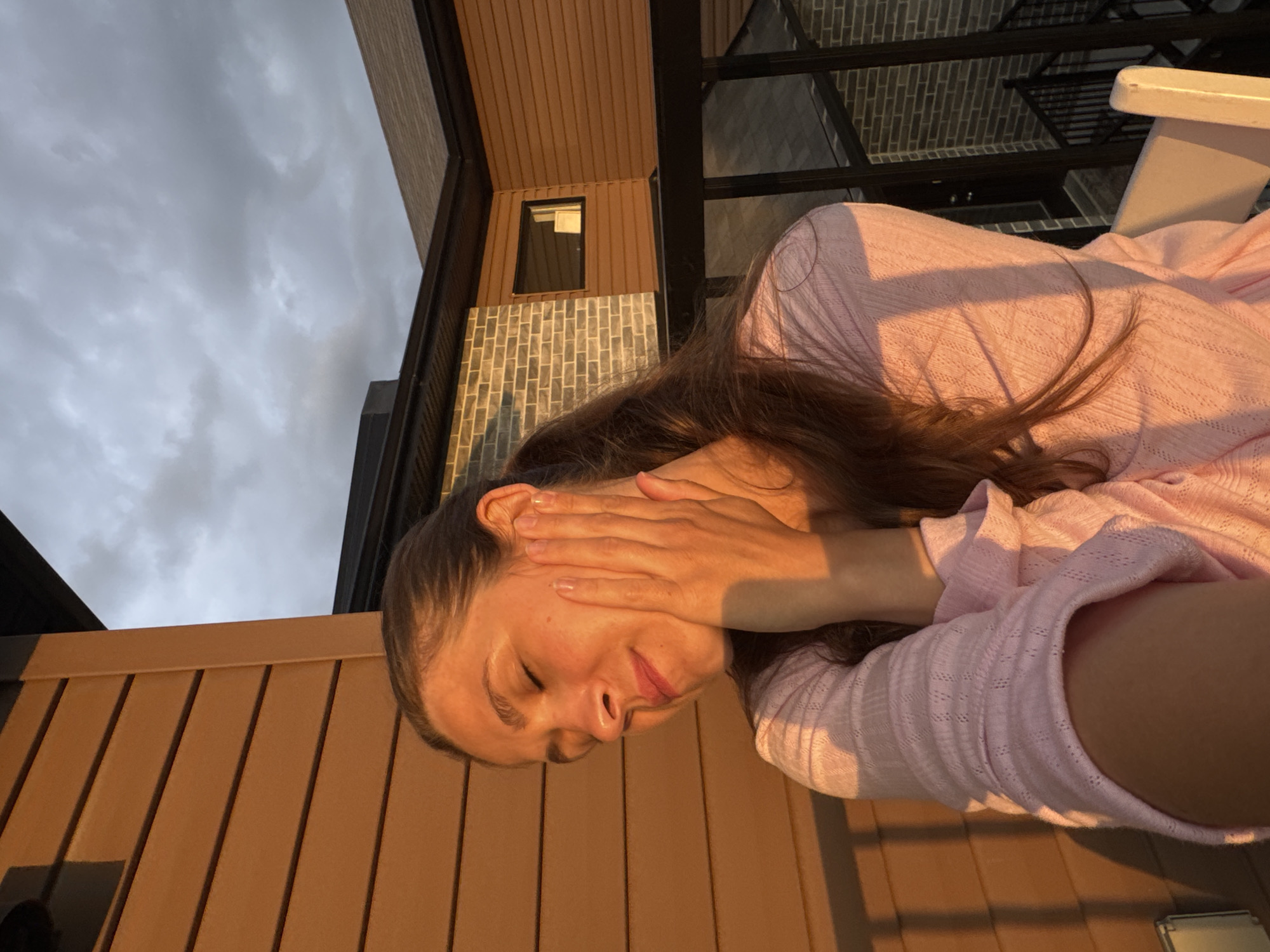 The image shows a woman with her eyes closed, holding her hand to her face, bathed in warm sunset light. She is wearing a pink knitted top. Behind her, a modern building with brown and grey brick siding is visible. The sky is cloudy with hints of grey and white. The scene appears to be on a balcony or patio. This location is in Gatineau, Canada.