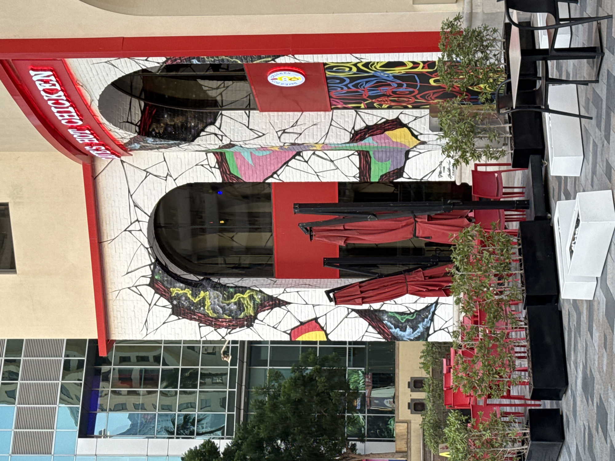 The image shows the exterior of a restaurant named "Dave's Hot Chicken" in Dubai, United Arab Emirates. The restaurant's facade is decorated with a white brick pattern that appears cracked, revealing vibrant graffiti underneath. Red awnings are visible over the arched doorways. In front of the restaurant, there is an outdoor seating area with red chairs and tables. Several black planters filled with green shrubs are placed in front of the seating area. To the left of the restaurant, a modern glass-fronted building reflects the surrounding environment. The ground is paved with grey and white tiles. No people are visible in the image.