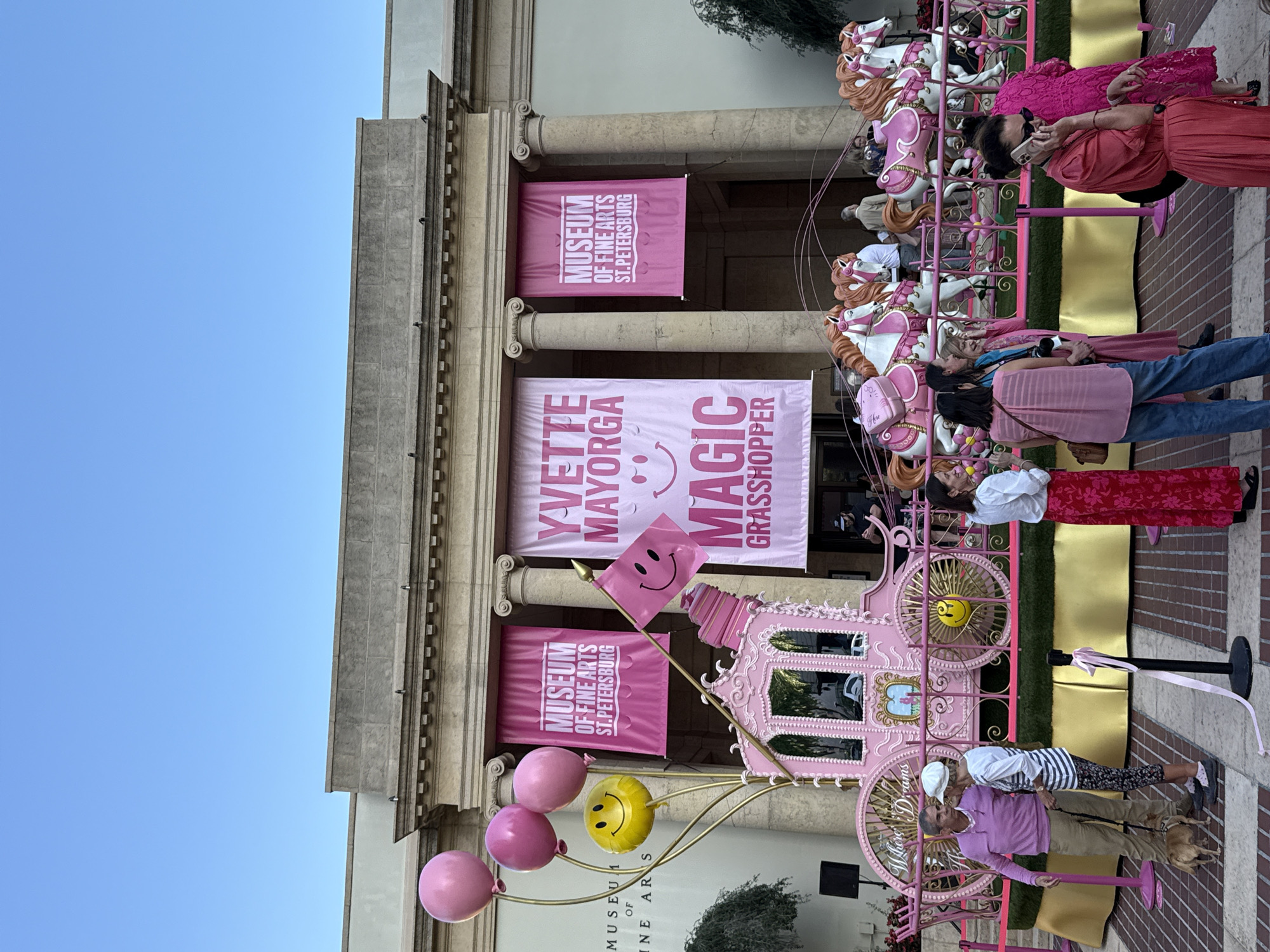 An outdoor scene is depicted in front of the Museum of Fine Arts, St. Petersburg, United States of America. A large, ornate pink Cinderella-style carriage, embellished with gold accents and a smiley face on a wheel, stands prominently on a raised platform. The carriage is partially labeled "Where Dreams." From its left side, a cluster of three pink balloons and one yellow smiley-face balloon rises on gold stalks. A pink flag with a smiley face is affixed to the carriage's rear. In front of the carriage, on the same platform, several white and pink carousel-style horses are arranged as a team. The platform is covered in green artificial turf with a gold-colored skirting, and its perimeter is marked by pink ribbon barriers.
The museum building, characterized by its classical architecture with columns, serves as the backdrop. Three large pink banners hang from the facade: two outer banners display "MUSEUM OF FINE ARTS ST. PETERSBURG," and the central banner reads "YVETTE MAYORGA MAGIC GRASSHOPPER" with a large smiley face. The sky above is clear and blue.
Numerous individuals are present on the paved area in the foreground. On the left, a man in a pink sweater and khaki pants stands conversing with a woman in a white and black striped top and patterned leggings, holding a leash for a small, light brown dog. To the right, a woman in a long red dress is speaking on a mobile phone, while another woman in a pink top and blue jeans stands facing right. Other people are visible throughout the scene, observing the display or engaged in conversation. Many individuals are dressed in various shades of pink and red.