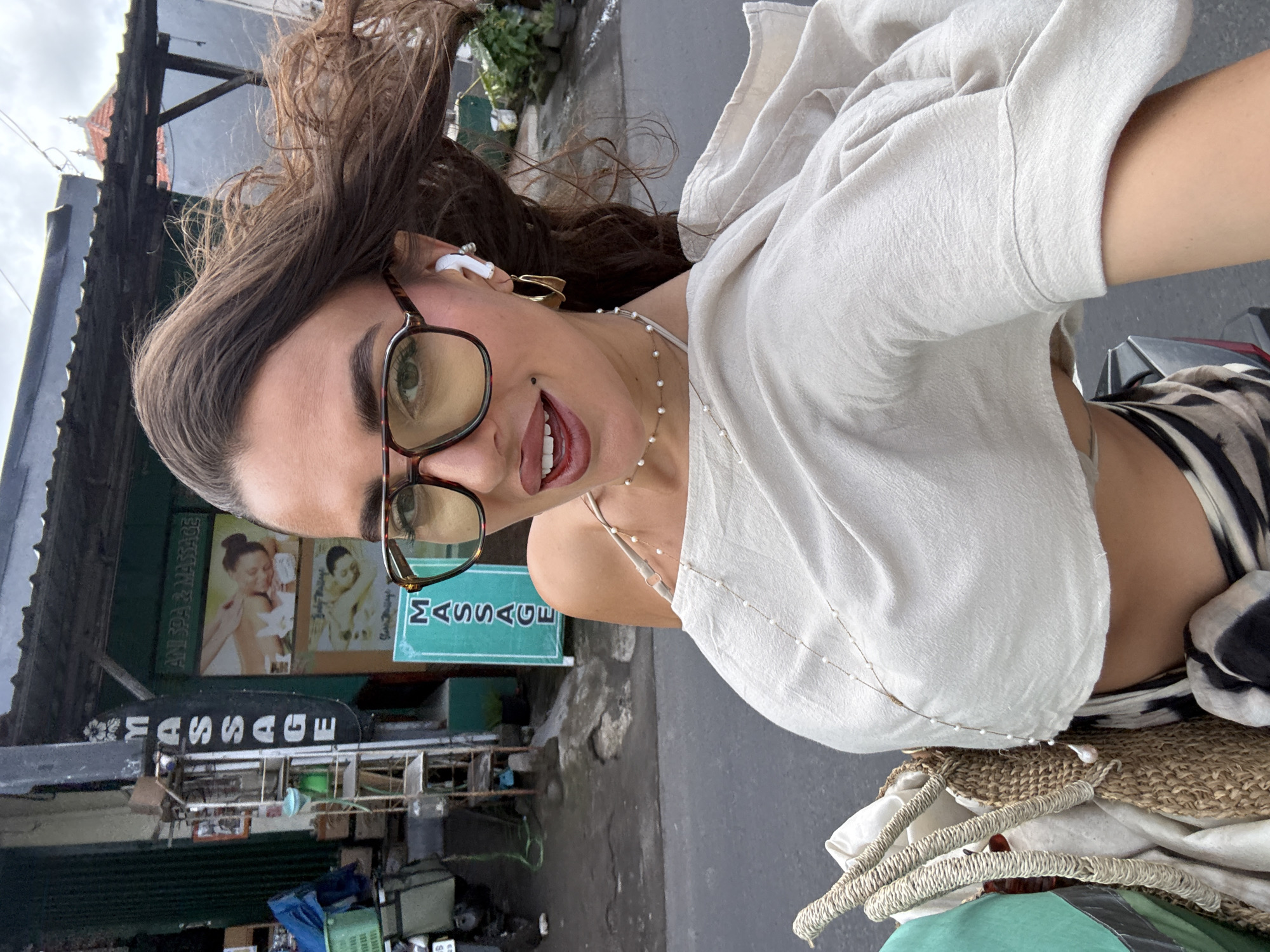 The image shows a woman with long brown hair, wearing glasses, a white shirt, and a pearl necklace, possibly taking a selfie. She is standing outside a building with a sign that says "MASSAGE," indicating a massage parlor. The surroundings appear to be a street scene in Kuta Selatan, Indonesia, with typical Balinese architecture, signs, and urban elements. A motorbike is partly visible on the right side of the image, and what seems to be a straw bag is on top of it.