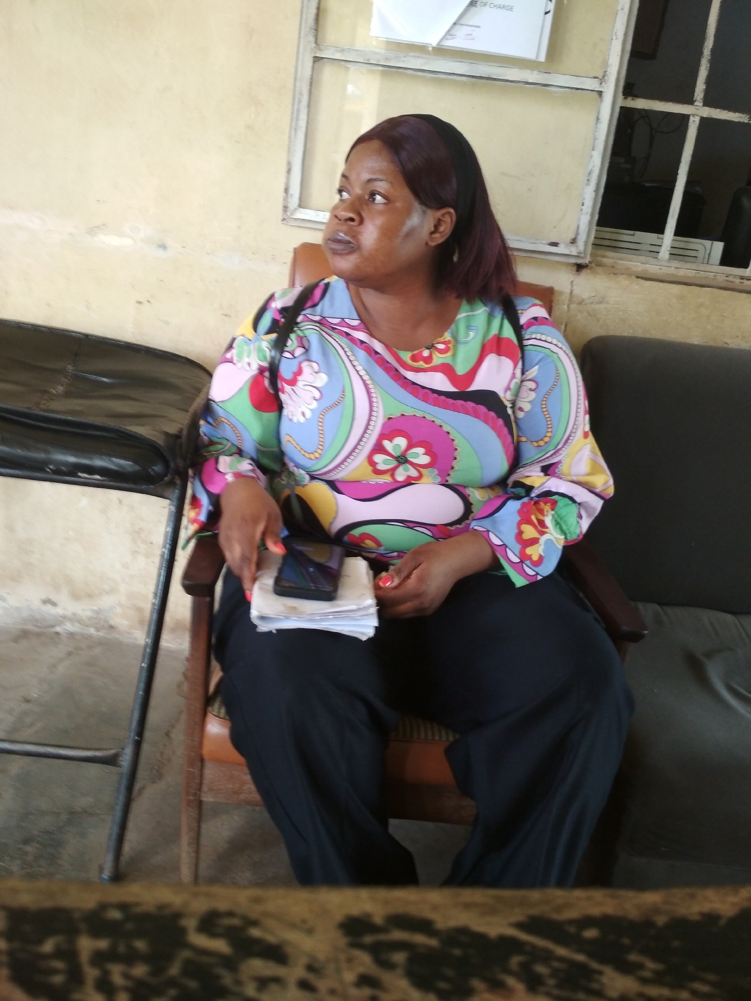 A medium shot captures a Black woman sitting in a brown wooden chair in what appears to be an office or waiting area. She is facing to her left, looking off-camera. She has dark skin and dark hair, with a maroon-colored headband. She is wearing a brightly colored, long-sleeved blouse with a paisley pattern in shades of pink, blue, green, and yellow, and dark pants. Her right hand is resting on a stack of papers, and a dark-colored smartphone is placed on top of the papers. To her left is a black metal chair with a black leather seat. Behind her, a window with metal bars is visible, with a document pinned to the wall above it. The walls are a pale yellow. The floor is concrete. The scene is likely within a building in Bisichi, Nigeria, given the context of the question.