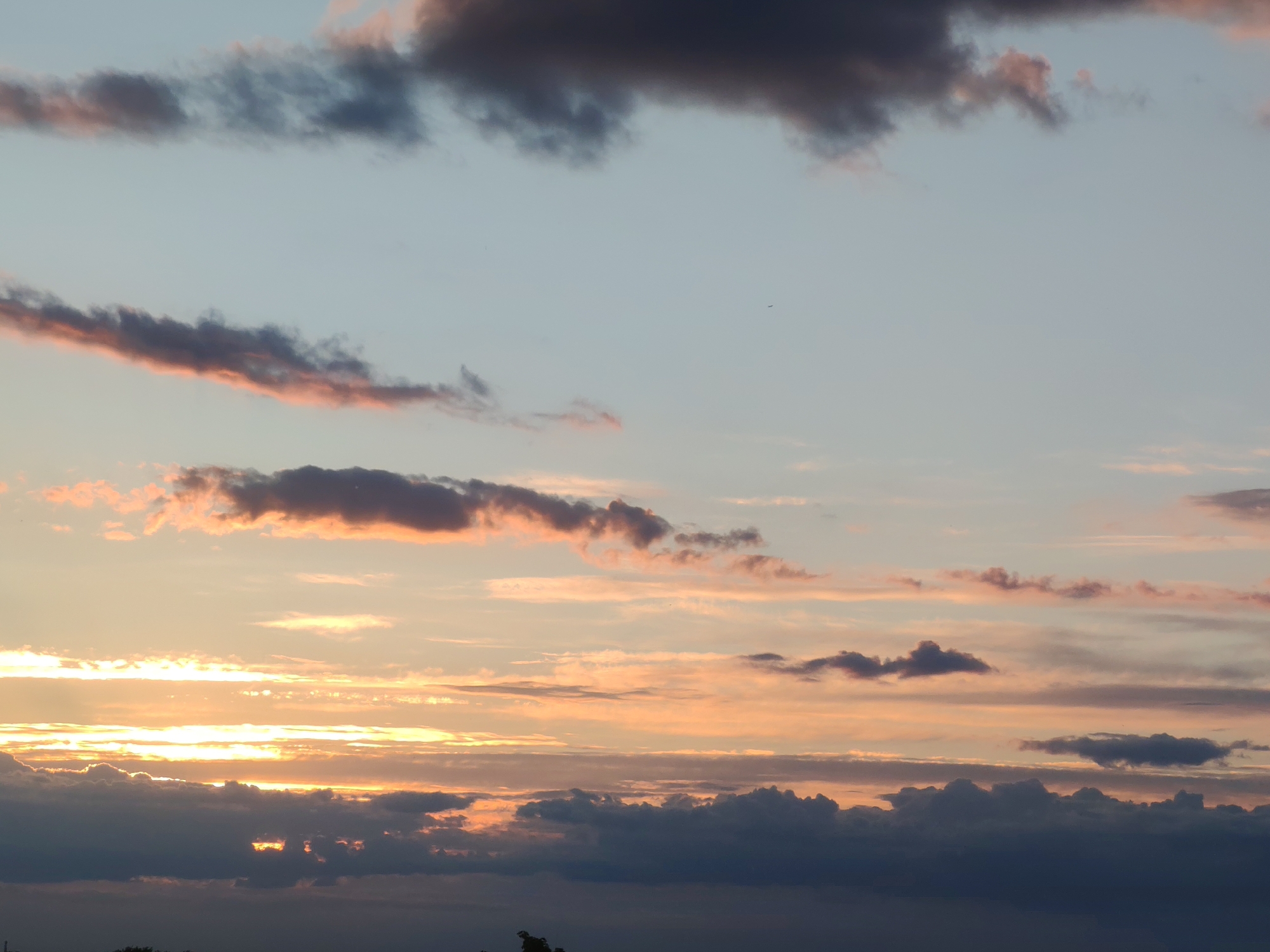 The image displays a vast expanse of sky dominated by numerous clouds during either a sunrise or a sunset, as indicated by the warm, golden light. The scene is set against the backdrop of Podolsk, Russia, although no specific location cues are visible within the image itself.
The upper portion of the sky is a soft, muted blue-grey, traversed by long, dark, horizontal cloud formations with hints of pink and purple where they catch the ambient light. Below these, the sky gradually transitions into warmer tones, with light blues and faint pinks giving way to vibrant oranges and yellows closer to the horizon.
The middle and lower sections of the frame are particularly striking, showcasing several layers of clouds intensely illuminated by the sun's low angle. Bands of stratiform clouds glow brightly with golden and orange light, particularly towards the left where the light source seems most intense. Some clouds appear thin and stretched, allowing light to diffuse through them, while others are denser and more textured, creating silhouettes against the bright background. Towards the bottom of the image, heavy, dark grey and bluish cloud masses form a substantial base, with glimpses of brilliant light breaking through their denser parts.
No people or distinct man-made objects are visible. A very small, dark speck, possibly a bird or a distant aircraft, can be observed in the upper-middle section of the sky. The overall atmosphere conveyed is one of natural beauty and serene transition as day either begins or ends. There is no visible text in the media file.