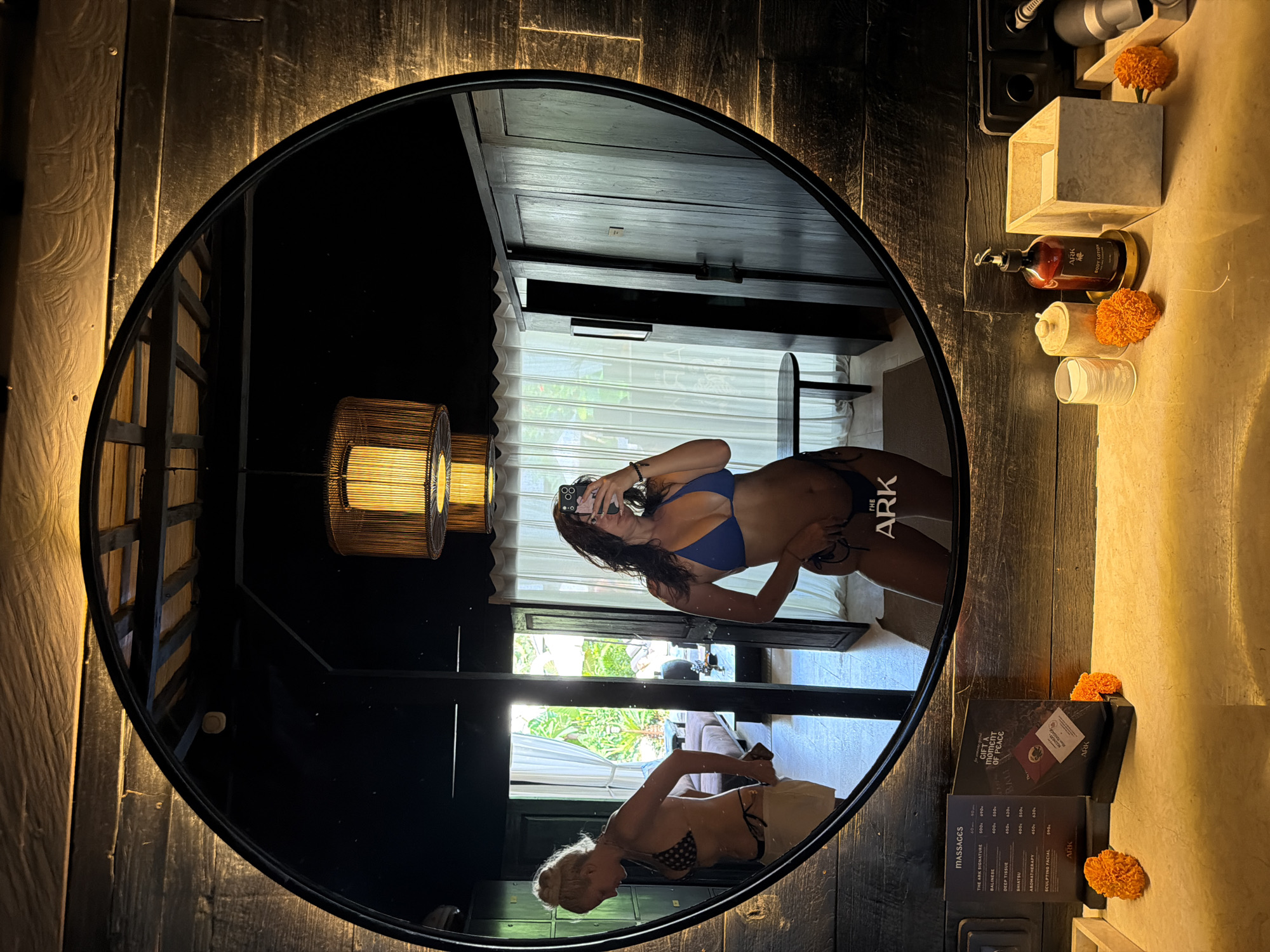 In a bathroom with dark wooden walls and a large circular mirror, two women are visible in the reflection. The woman on the right is taking a selfie, wearing a blue bikini. The woman on the left is facing away from the camera, adjusting her bikini bottoms, wearing a polka dot bikini top and white shorts. The room appears to be a resort in Mengwi, Indonesia, as indicated by the "THE ARK" logo on a menu in the foreground. The bathroom counter holds various items including bottles of lotion and a tissue holder.