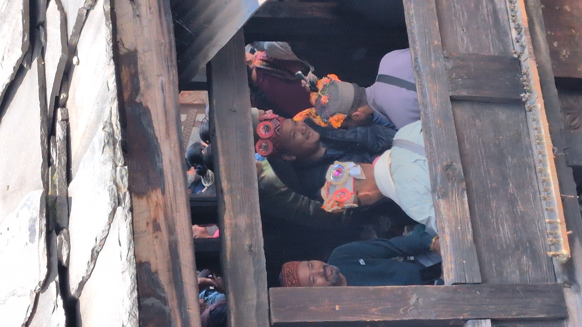 A medium-shot, eye-level, vertical image shows a group of men in traditional Himachali hats standing on a wooden balcony in Malana, India, likely during a festival. The men are gathered close together and appear to be engaged in conversation. The man on the far left is wearing a red and black patterned cap, a dark green long-sleeved shirt, and has a beard. To his right, a man with a red hat adorned with circular, dark red embellishments and bright pink accents is smiling and looking to his left. In the foreground, a man in a light-colored garment and a white hat decorated with colorful pompoms and sequins is facing away from the camera, looking towards the man in the red hat. Behind him, another man wears a grey cap with orange floral decorations and a light purple shirt. The background is dark, suggesting an interior space or a shaded area. The balcony is constructed of rough-hewn wooden planks, and above them, the distinctive slate roofs of Malana are visible, characteristic of its unique architecture. The lighting is bright, indicating daytime.