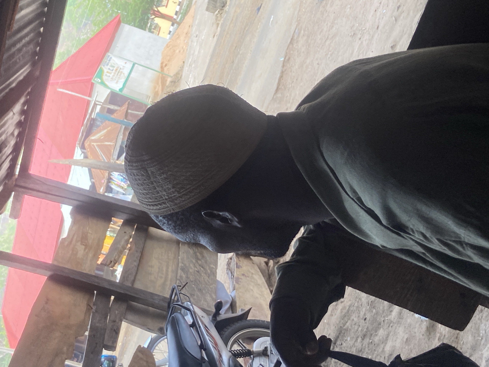 A man is depicted in a side profile, seated or bending within a shaded, rustic structure. He wears a dark, patterned kufi cap and a dark-colored long-sleeved garment. His head is bowed, and he appears focused on an unseen task or object located below the frame, with his right arm partially visible.
The immediate surroundings consist of rough wooden beams and planks, suggesting an open-sided shelter or workshop, with a corrugated metal sheet forming part of the left side. Through the openings of this structure, an outdoor scene is visible.
In the background, a reddish-pink building with a sloped roof stands prominently. Next to it, a light-colored sign with green accents displays the letters "C.R.C.K" along with other illegible text. Further behind, another building with a yellow wall and a darker roof can be seen. The ground outside appears sandy and unpaved. Parts of a motorcycle or scooter, including a wheel and handlebars, are partially visible in the lower foreground.
The lighting indicates it is daytime, with the outdoor areas brighter than the shaded interior of the shelter. The setting suggests an urban or semi-urban environment in Mararaba, Nigeria. The man's expression is not discernible due to the angle and shadows, but he appears to be concentrated on his activity.