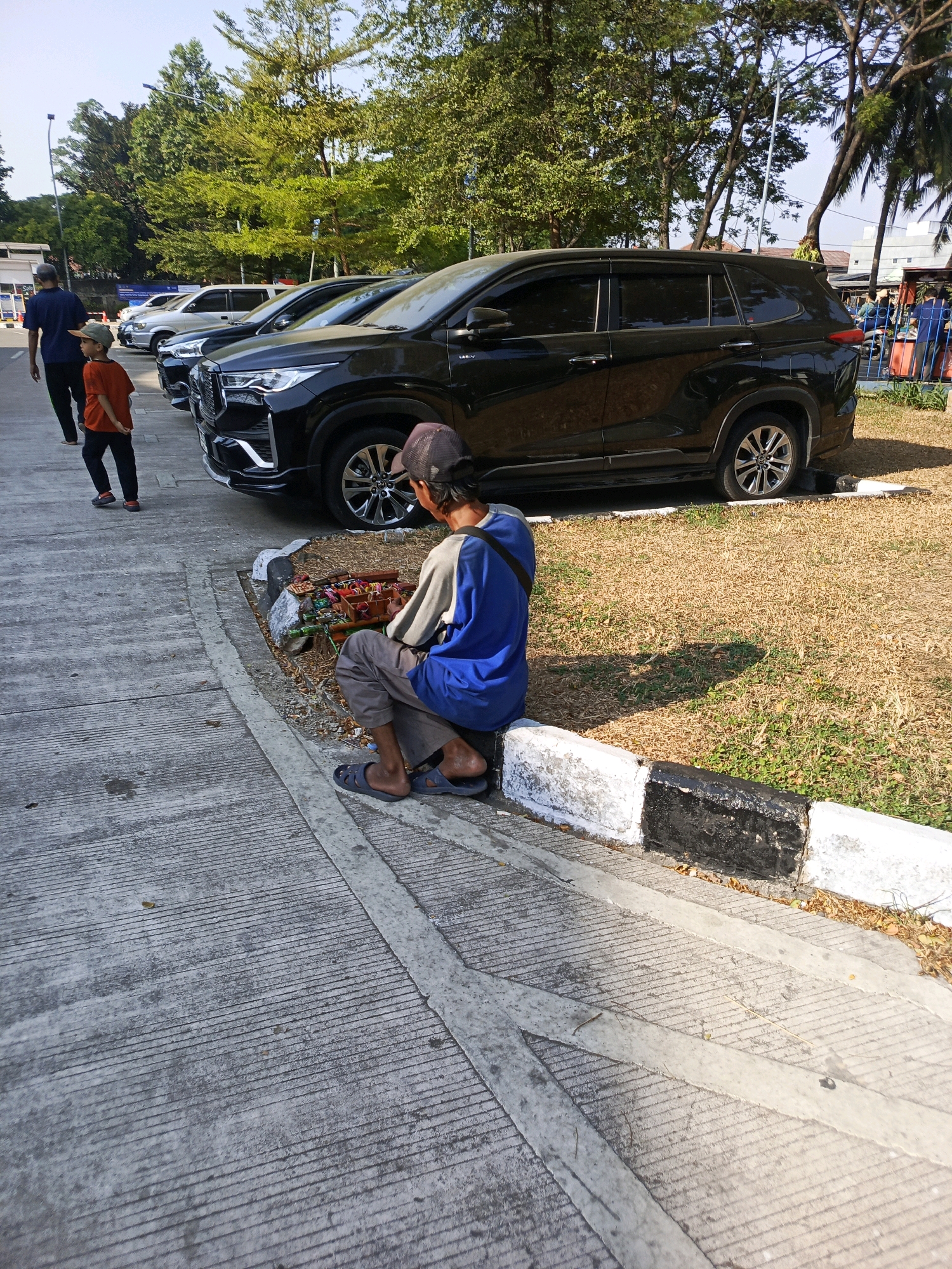In Tangerang, Indonesia, a man in a blue and gray shirt and a cap sits on a curb next to a black SUV. He is facing away from the camera, towards a collection of items on the ground that appear to be small goods for sale. To his left, a boy in an orange shirt and a man in a dark blue shirt and a cap walk away from the camera along a concrete path. The scene takes place in a parking lot with a grassy area bordering the paved surface. Trees and buildings are visible in the background.