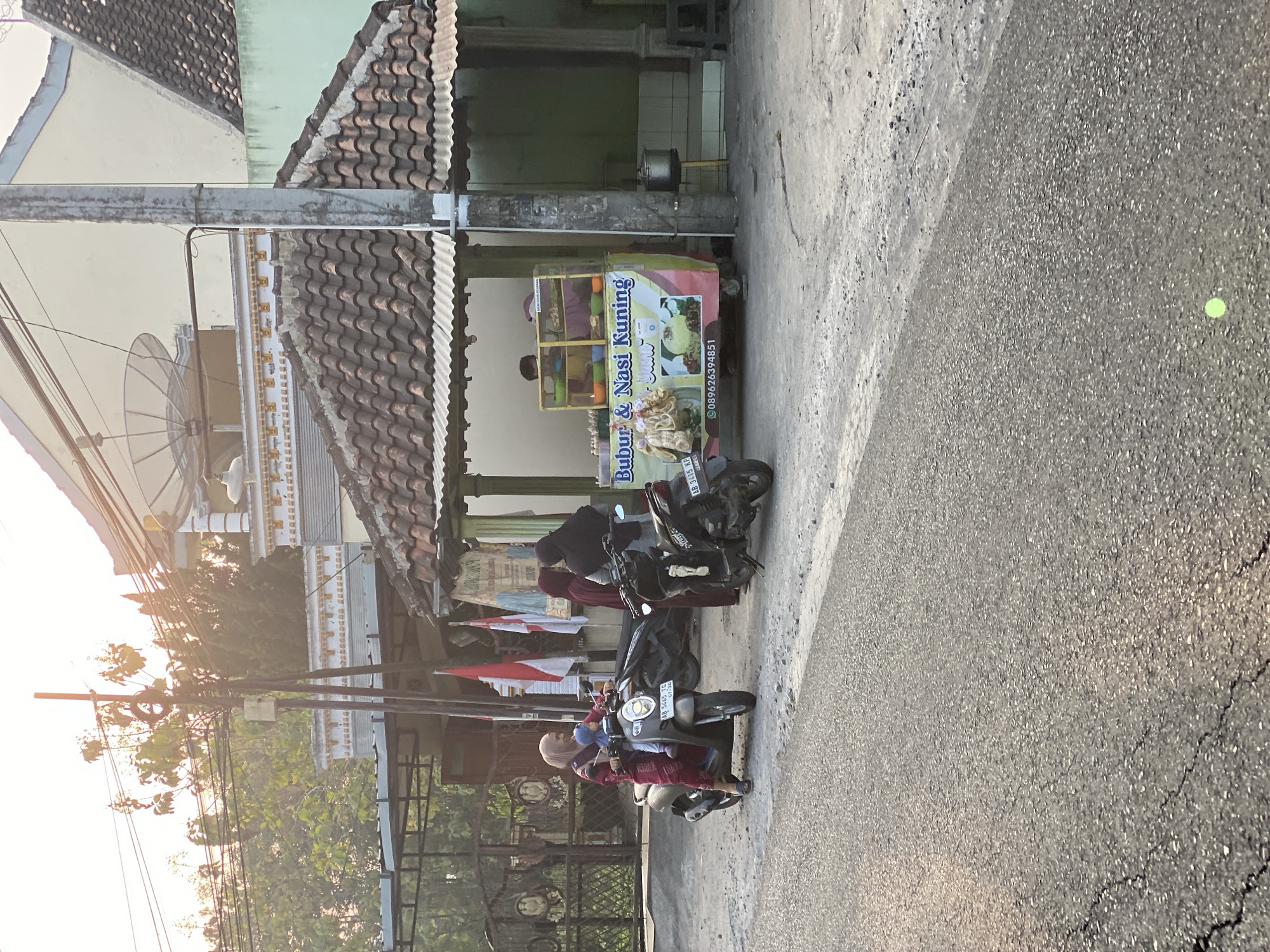 In Kasihan, Indonesia, at what appears to be a roadside stall, a woman wearing a maroon hijab and a matching dress is seated on a scooter with a young child in a blue hijab also on the scooter. The woman is looking to her left. Behind them, two other women, one in a dark maroon hijab and the other in a black hijab, stand near a food stall. The stall features a colorful banner advertising "Bubur & Nasi Kuning" and displays food items. Several motorbikes are parked near the stall. The scene is set on a paved road with a cracked surface, with buildings and trees in the background. The sun is low in the sky, casting long shadows and creating a warm, golden light.