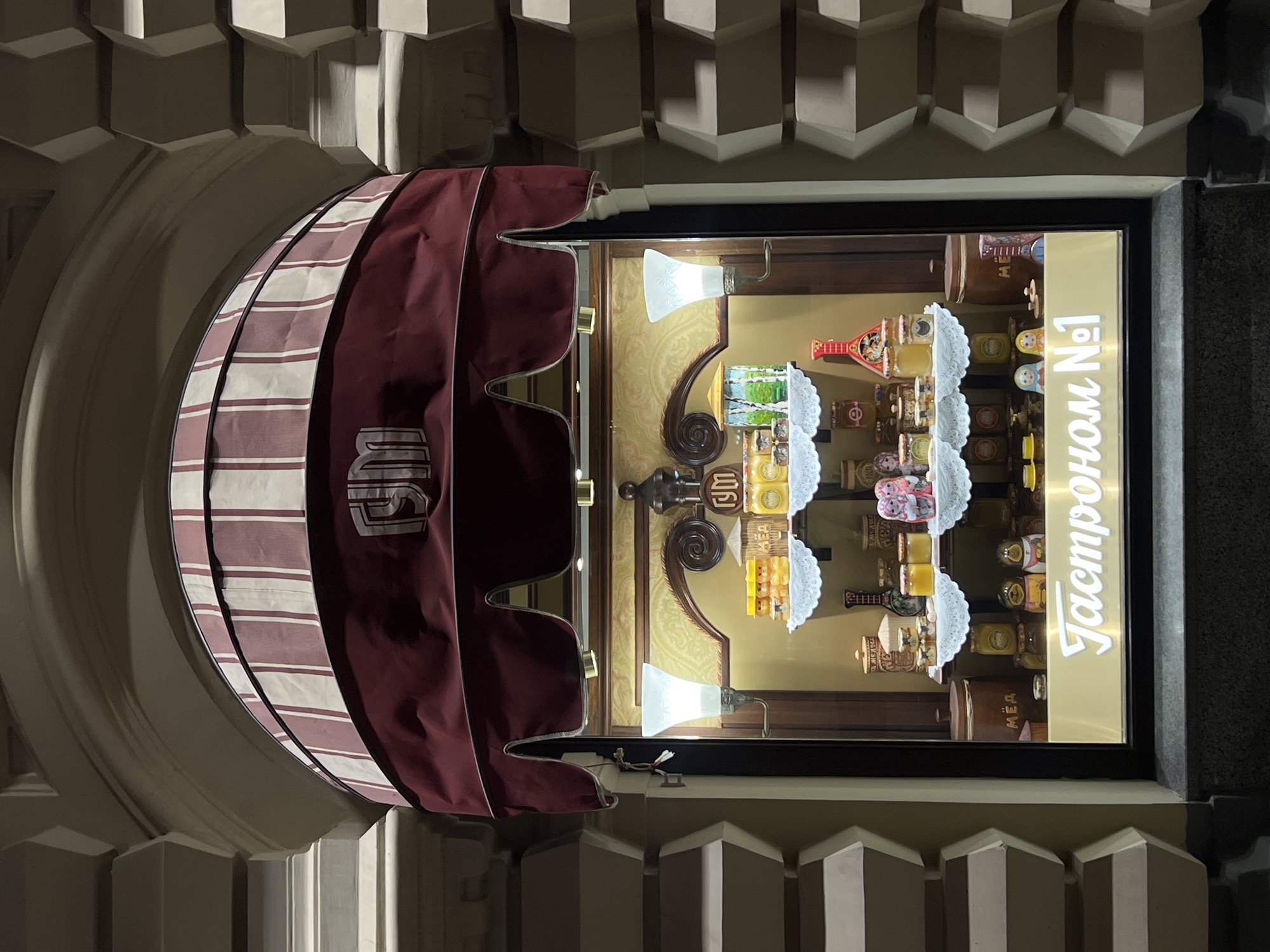 A storefront in Moscow, Russia, features a window display with a red and white striped awning above. The window showcases various items, including jars of honey, painted wooden dolls, and decorative objects on shelves and platforms. Lamps illuminate the display. The store's name, "Gastronom No 1", is visible in a vertical, illuminated sign. The building's facade has a decorative, columned design.