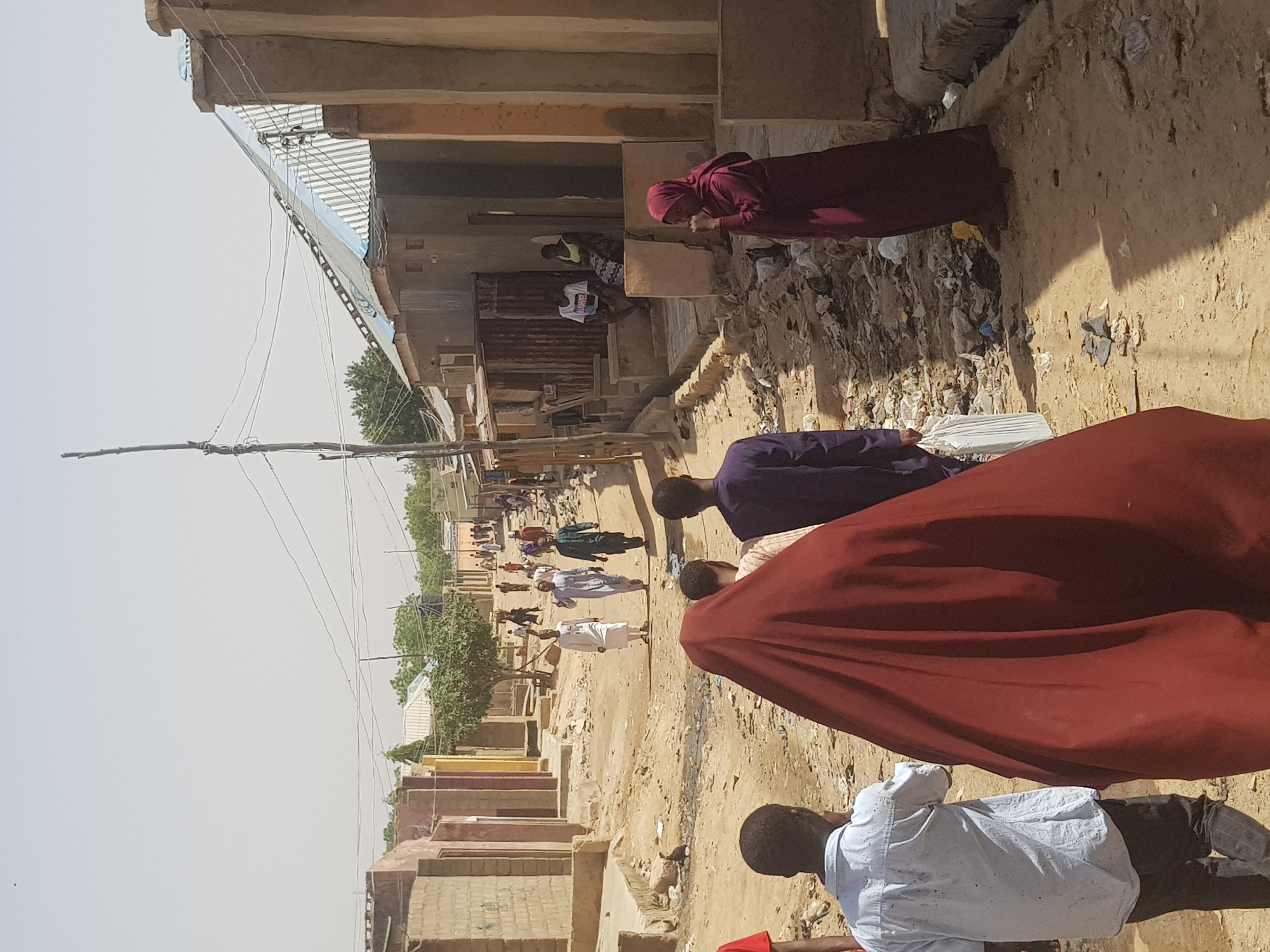 The image depicts a dusty street scene in what is possibly Dabawa, Nigeria. The street is lined with buildings, some with corrugated iron roofs. Several people are walking along the street, and others are visible in front of the buildings. Debris is visible alongside the buildings in the foreground. A woman in a red hijab is standing. Overhead powerlines stretch across the sky. The foreground is dominated by an individual partially blocking the camera.