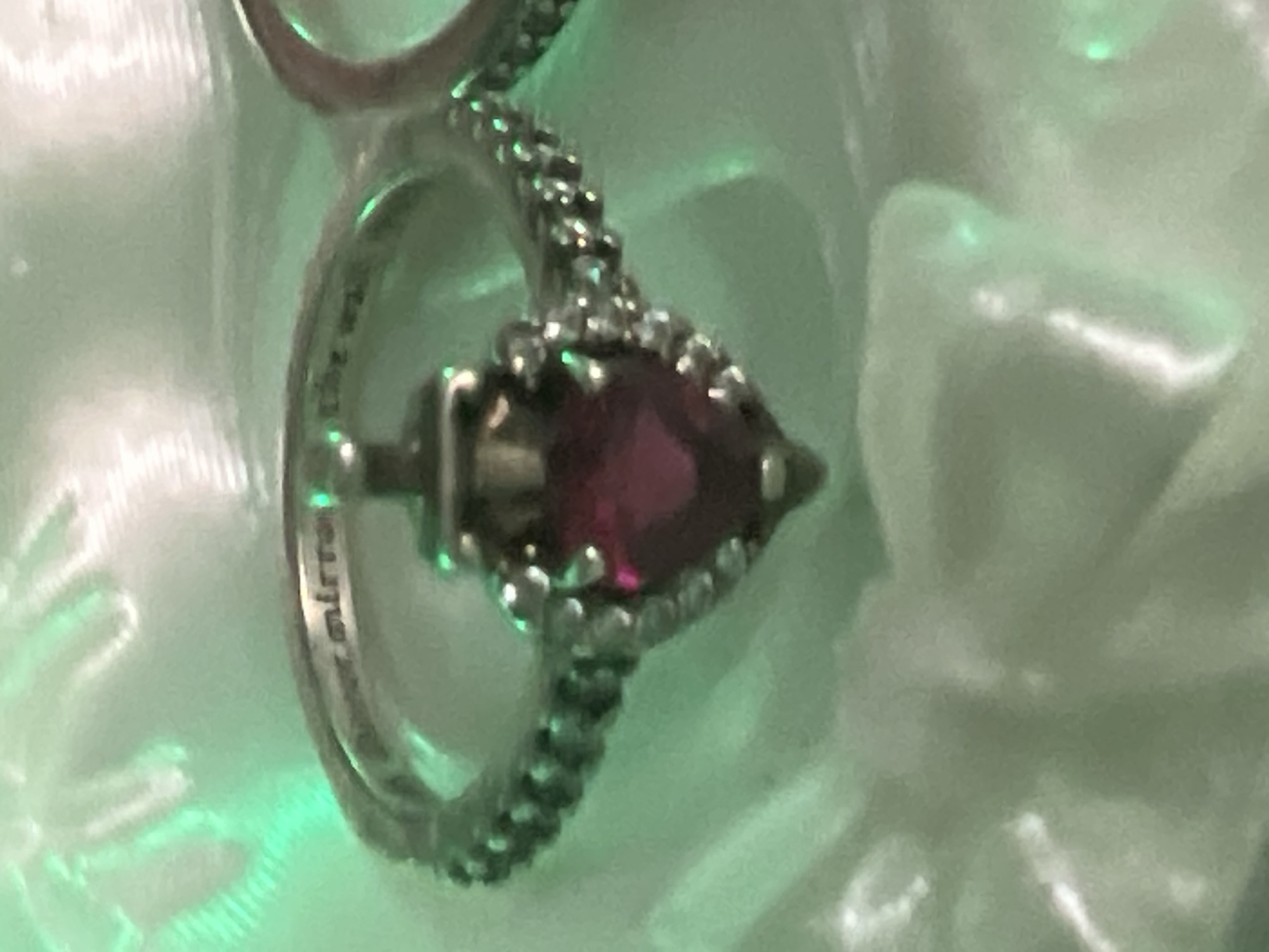 The image displays a close-up view of a ring. The ring is silver and features a heart-shaped red stone in the center, surrounded by smaller clear stones. Engraved text is visible on the inside of the band, which appears to read "never mirror the wall". The background is a blurry, light-colored fabric, possibly a tablecloth or garment, with green lighting casting shadows and reflections. The overall aesthetic suggests a personal item, perhaps on display or being worn. The context of the city, state, or any specific location is not discernible from the visual information provided.