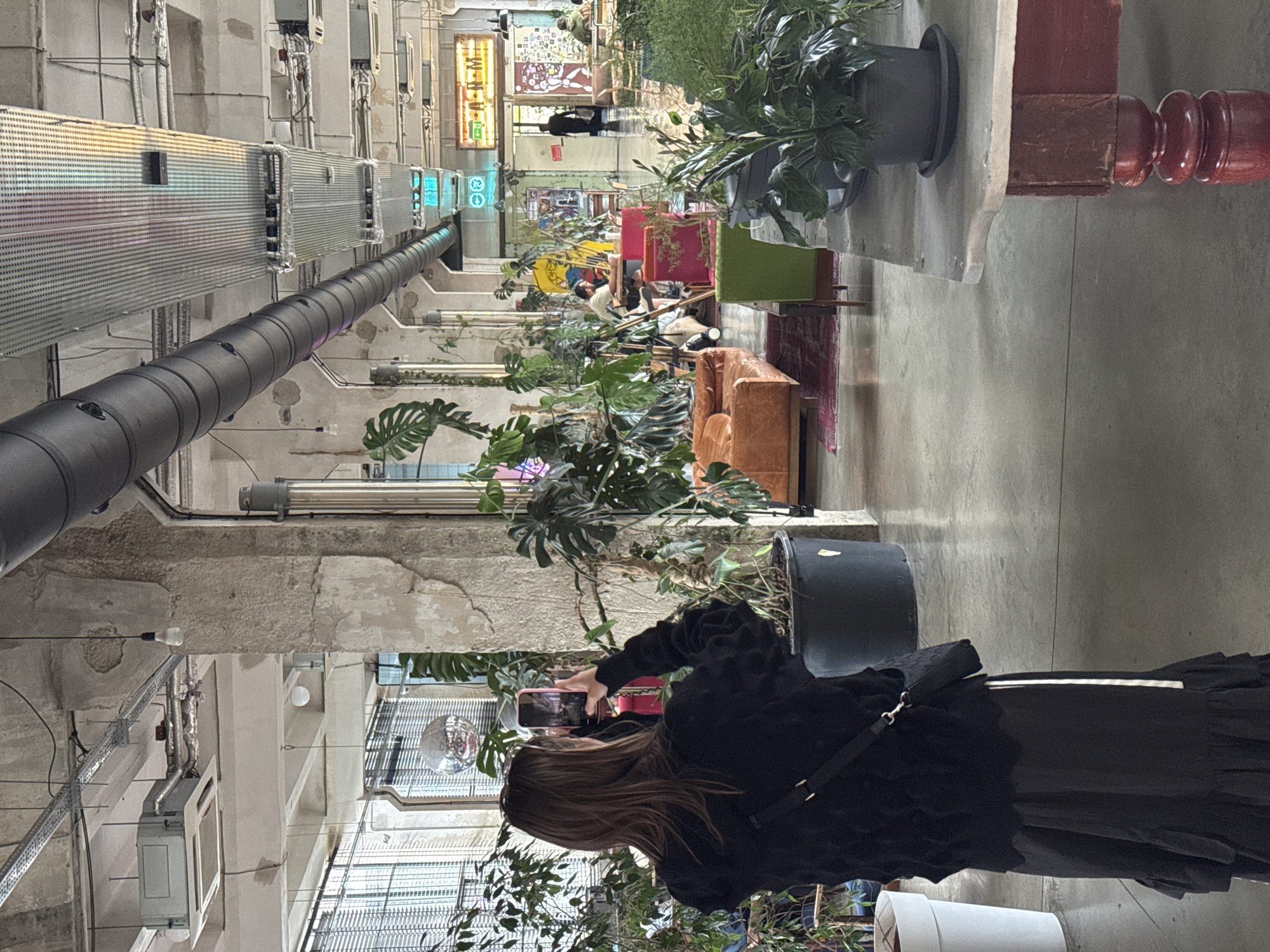 The image depicts the interior of a large, industrial-style space with exposed concrete ceilings, pipes, and columns, characteristic of urban loft or repurposed factory settings. The space is filled with abundant greenery, with numerous potted plants of various sizes and types contributing to a biophilic atmosphere.
In the foreground, a person with long brown hair, seen from behind, is wearing a black knitted sweater and a long black skirt. They are holding a smartphone in their right hand, appearing to take a photograph or video of the scene. A black crossbody bag with a white strap detail is worn over their shoulder.
The environment beyond the foreground includes a variety of seating arrangements, suggesting a communal or co-working space, cafe, or exhibition area. A burnt orange leather sofa is visible, along with a bright green chair. Further into the space, there are tables and chairs where other individuals are seated and interacting. One person is seated at a table, appearing to be working or engaged in conversation. In the background, a person is walking away from the camera towards a doorway.
Signage with neon lighting is present in the distance, one appearing to be a yellow and orange sign with text, and another with green lettering. The overall impression is of a trendy, contemporary urban venue, potentially a cultural hub or a stylish cafe located in Tbilisi, Georgia, given the distinctive architectural style often found in the city's renovated industrial zones.