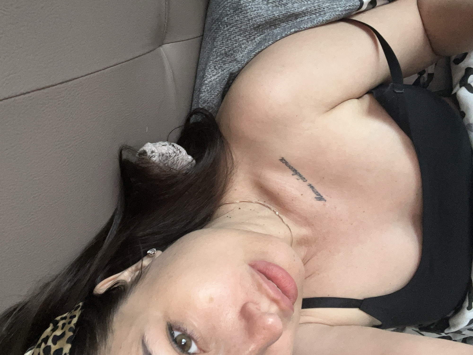 A woman is lying down, facing the camera, with her head tilted to the side. She has dark hair, styled with a leopard-print headband, and brown eyes. She is wearing a black tank top, and her left shoulder is exposed. A tattoo is visible on her upper chest. A gray knitted blanket is draped over her shoulder and back. The background appears to be a textured surface, possibly a couch. The lighting is soft and natural. The image is a close-up shot, focusing on the woman's face and upper body.