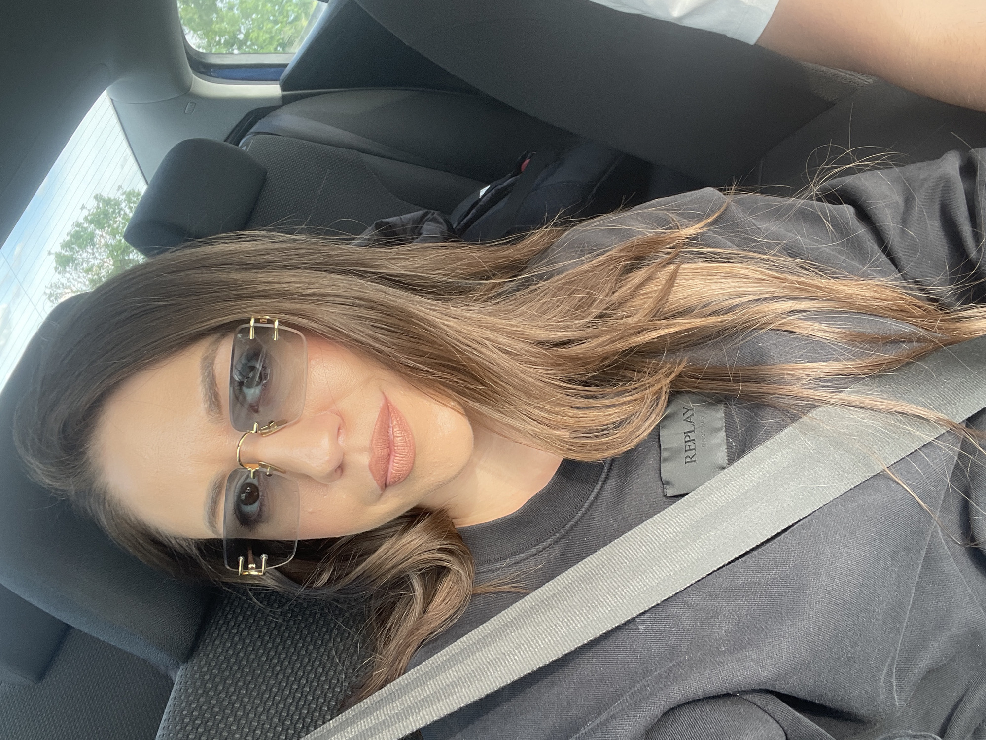 A woman with long brown hair and sunglasses lies reclined in a car seat, wearing a black shirt with a gray seatbelt across her. The scene is inside a car, with the car seat and other interior parts visible. The location, Shishmantsi, Bulgaria, is not identifiable from the image alone.