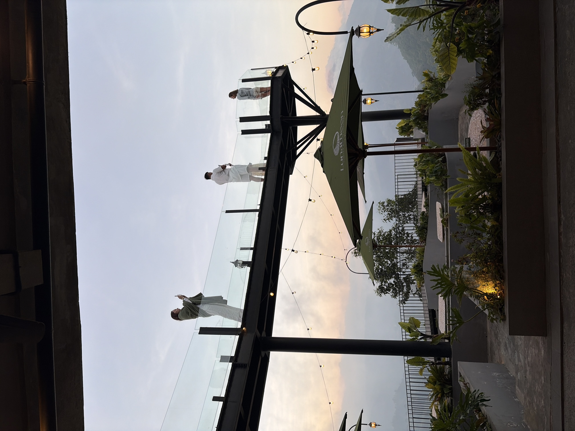 The image depicts an elevated outdoor seating area in Kintamani, Indonesia, offering a panoramic view of lush, mist-covered mountains. In the foreground, several green umbrellas are set up, with ornate lanterns hanging from curved metal poles. Planters filled with tropical foliage line the edges of the seating area. Above, a glass-railed walkway juts out, supported by dark metal beams. Three individuals are present on the walkway. On the left, a person in a green shawl and white pants stands with a phone, seemingly taking a photo. To the right, a person in white attire stands facing away from the viewer, also with a phone. A third person, dressed in a patterned top and shorts, stands to the far right, looking out at the view. The sky is overcast with a soft, diffused light, suggesting either early morning or late afternoon. The overall atmosphere is serene and scenic, highlighting the natural beauty of the Kintamani region.