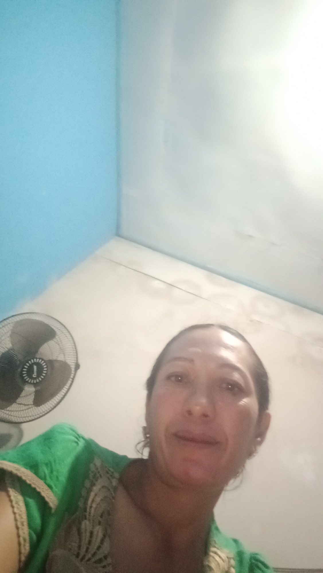 An indoor selfie shows a woman with dark hair and dark eyes, looking directly at the camera. She is wearing a green top with intricate gold detailing on the neckline. To her left, a black oscillating fan is visible, positioned against a light-colored wall. The background features a corner where a light blue wall meets a white wall, with a white ceiling above. The lighting is bright, suggesting the image was taken during the day or under strong artificial light. The scene appears to be a domestic setting, possibly a bedroom or living area, in Denpasar, Indonesia.