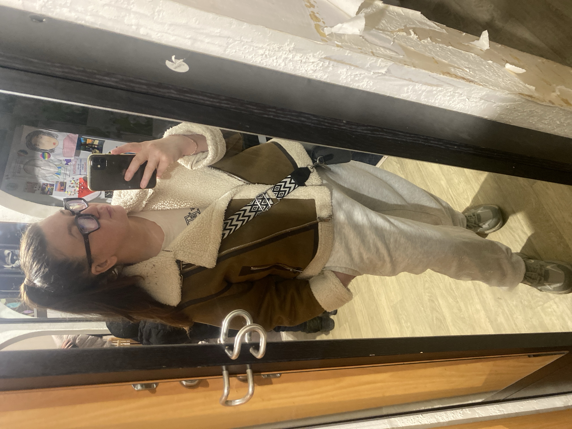 A woman is taking a selfie in a mirror. She is wearing glasses, a brown jacket with white fur trim, a white t-shirt, and grey sweatpants. The woman is holding a black smartphone. The mirror is above a wooden cabinet with metal handles. The background includes a wall with peeling paint and a wooden floor. The location is unknown.