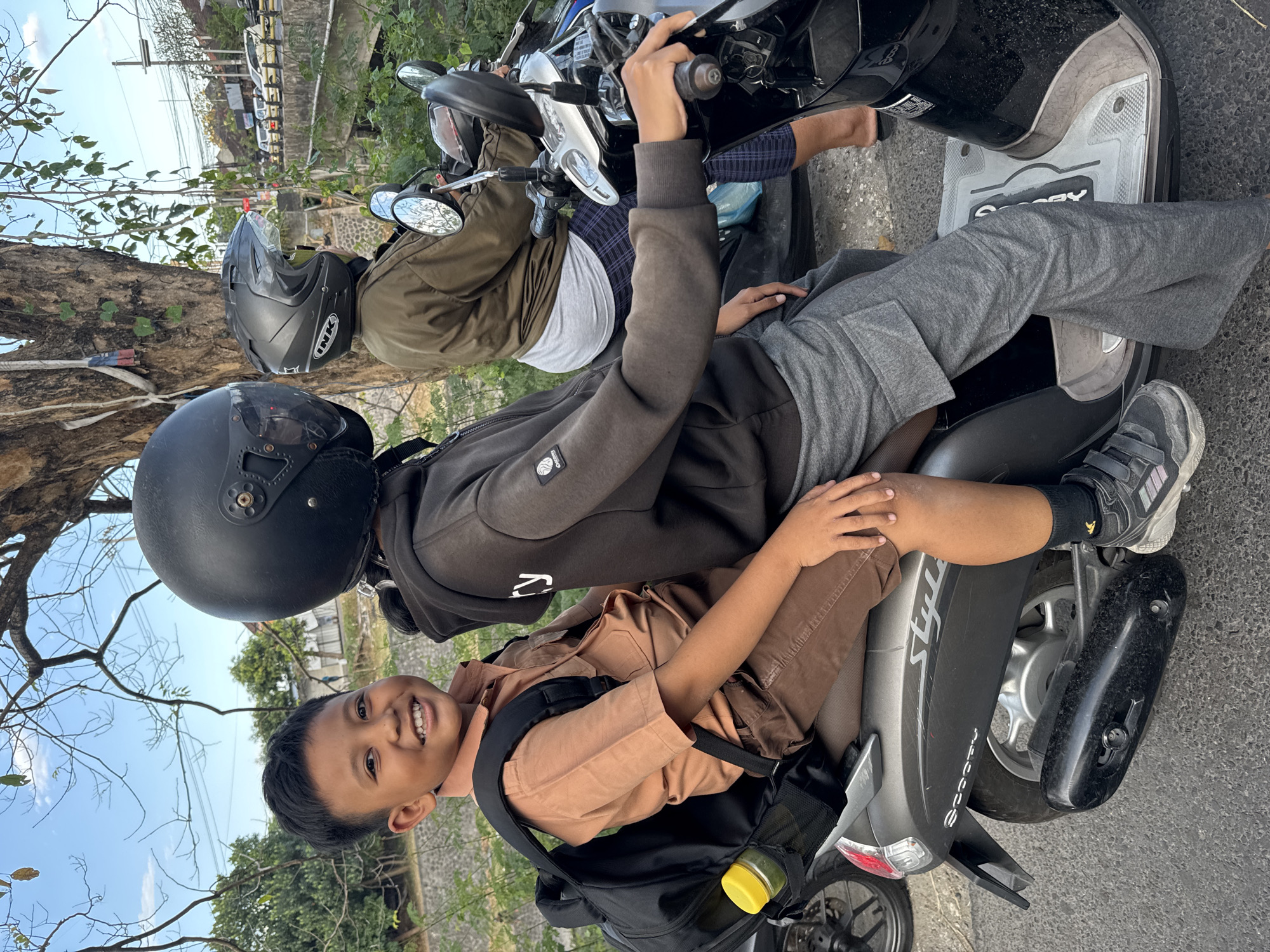 This is a medium shot, eye-level, outdoor photograph taken on a sunny day in Denpasar, Indonesia. Three people are on two scooters. In the foreground, a young boy wearing a tan school uniform and a black backpack is seated on the back of a scooter. He is smiling and looking at the camera. The scooter's make, "Scoopy," is visible on the rear fender. To his right, another person wearing a dark hooded sweatshirt and gray pants is seated on the same scooter, facing forward and wearing a black helmet. Their arm is extended, resting on the scooter's handlebars. Behind them, on a separate scooter, a third individual wearing a green jacket and plaid pants is visible from the shoulders up, also wearing a helmet. The background shows a tree with sparse leaves, a stone wall, dense green foliage, and a road with cars and power lines, indicative of an urban environment. The scooters are parked on an asphalt road.