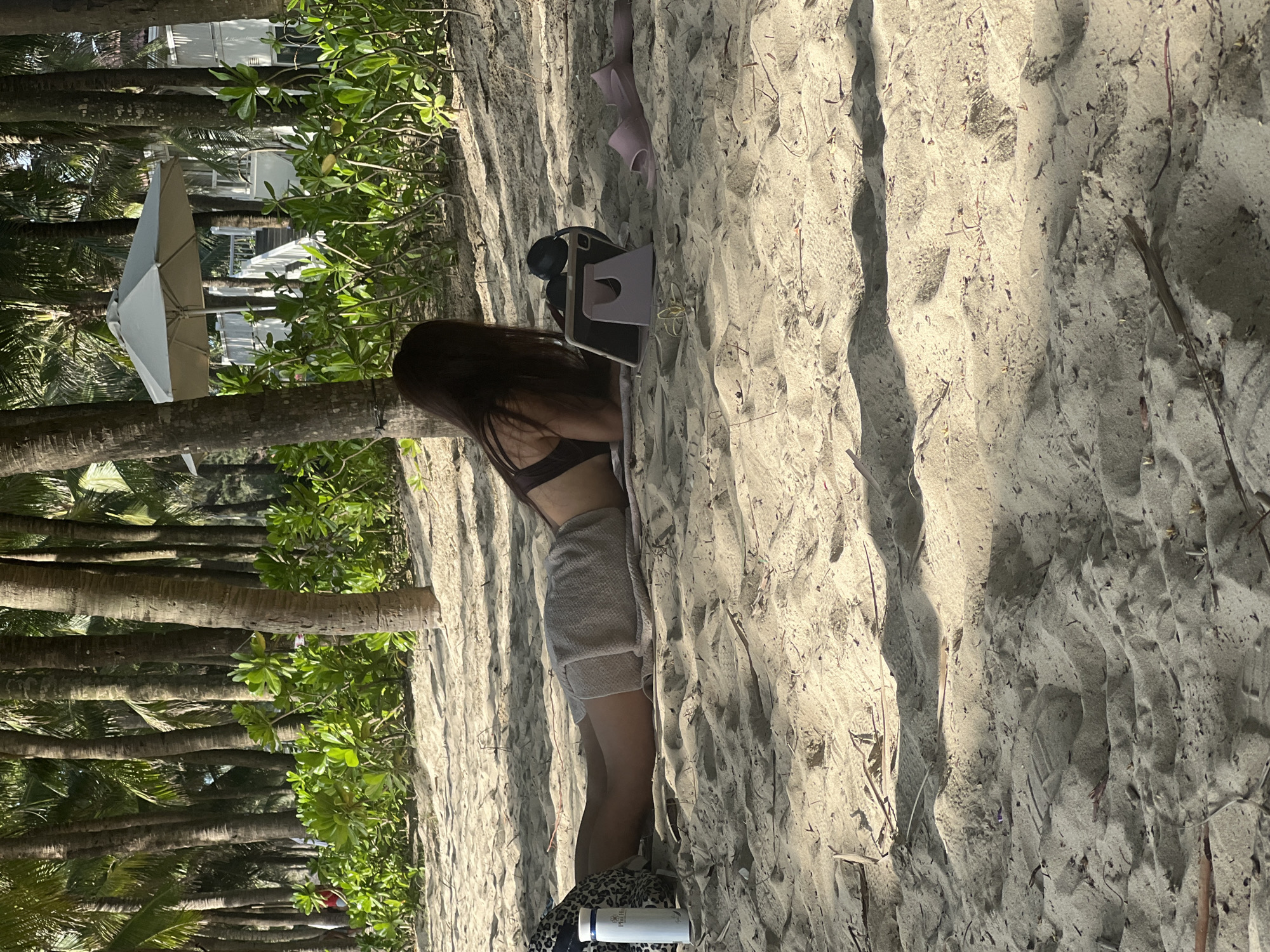 In Đà Nẵng, Vietnam, a person with long, dark hair lies on a sandy beach, facing away from the camera and towards a tablet device propped on a stand. They are wearing a dark bikini top and a light-colored skirt. To their left, a white water bottle sits near a patterned bag. In the background, a dense line of palm trees creates a tropical environment, with lush green foliage interspersed among the trunks. Further in the distance, a white umbrella shades a structure, suggesting a resort or hotel area. The sand is textured with shadows cast by the palm trees, indicating a sunny day. A pair of purple flip-flops lies to the right of the tablet.