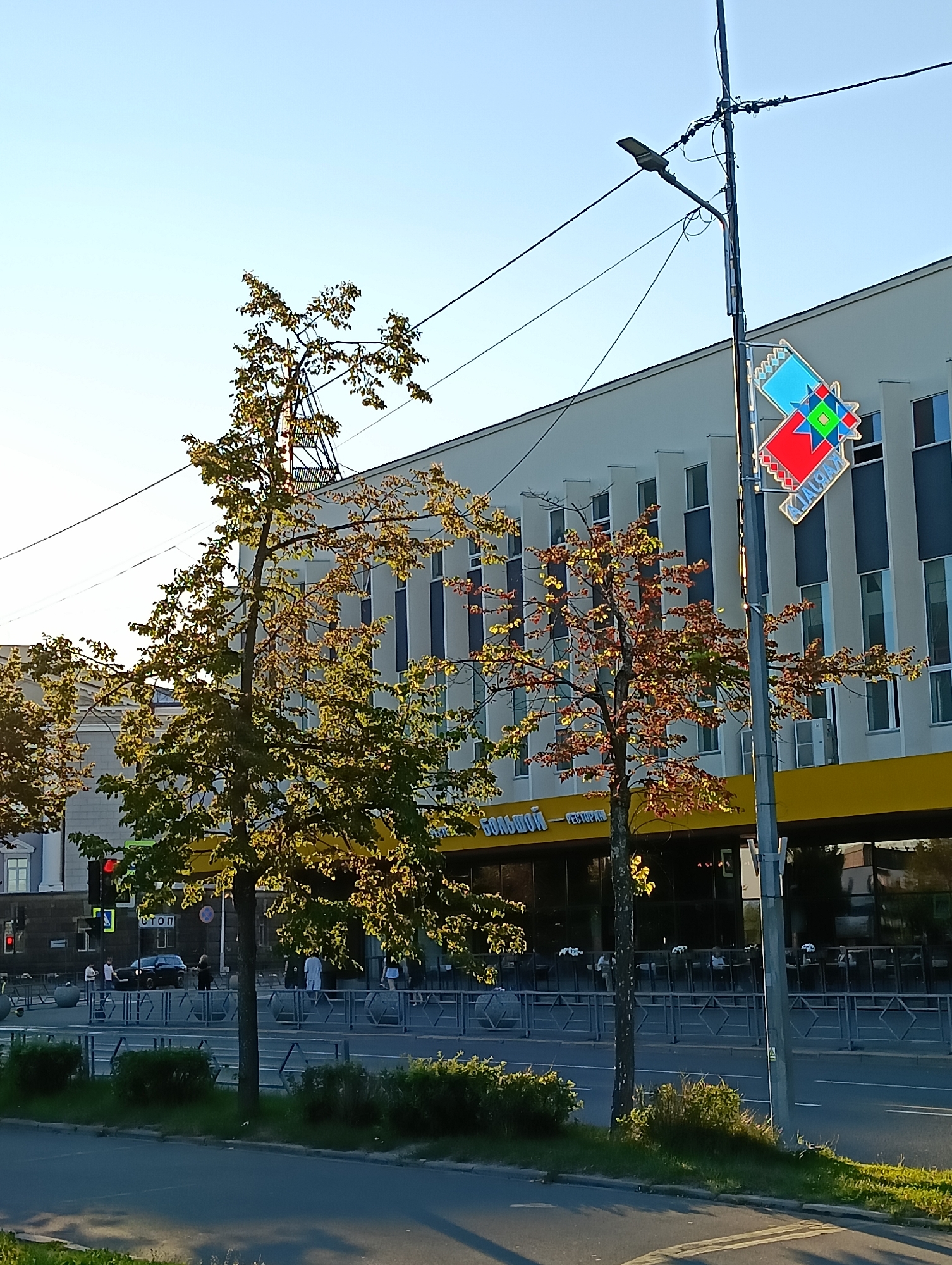 The image depicts a street scene in Petrozavodsk, Russia, likely during daylight hours given the bright sunlight. A modern building with a light-colored facade and dark-framed windows stands prominently. A vibrant yellow awning extends across the lower part of the building, and text above it is partially visible, with "БОЛЬШОЙ - РЕСТОРАН" (Bolshoi - Restaurant) discernible. Two trees are in the foreground, one with green leaves illuminated by the sun, and another with reddish-brown leaves, suggesting a transition into autumn. A street light with a modern fixture is attached to a utility pole on the right side of the image. A colorful, geometric sign, possibly a logo or decoration, is also mounted on the pole. The street in front of the building is lined with a metal fence, and several people are visible walking and standing, though their activities are not clearly discernible. A black car is parked near the building. The sky is clear and blue. The overall impression is a well-lit urban environment.