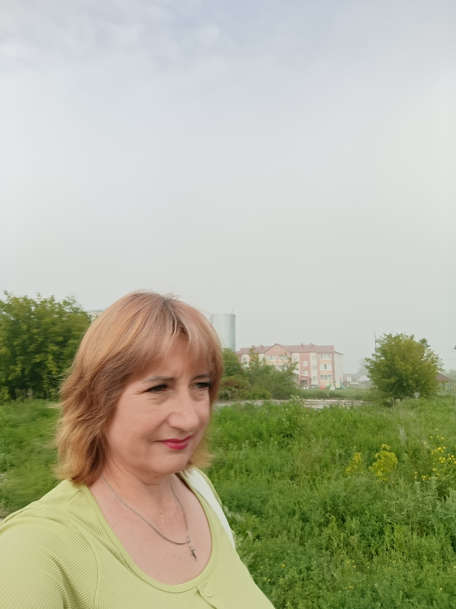 A woman with short, light brown hair is pictured in a close-up selfie. She is wearing a lime green shirt with a ribbed texture and a silver chain necklace with a small pendant. Her head is turned to the right, and she is looking away from the camera with a slight smile. Her lips are painted red. The background consists of a hazy sky, green grass, trees, and a building. The building appears to be a multi-story structure with a red roof and light-colored walls. The overall scene suggests an outdoor setting, possibly in a rural or suburban area of Tugulym, Russia, given the presence of the building and the natural landscape. The hazy conditions may indicate fog, mist, or dust in the air.