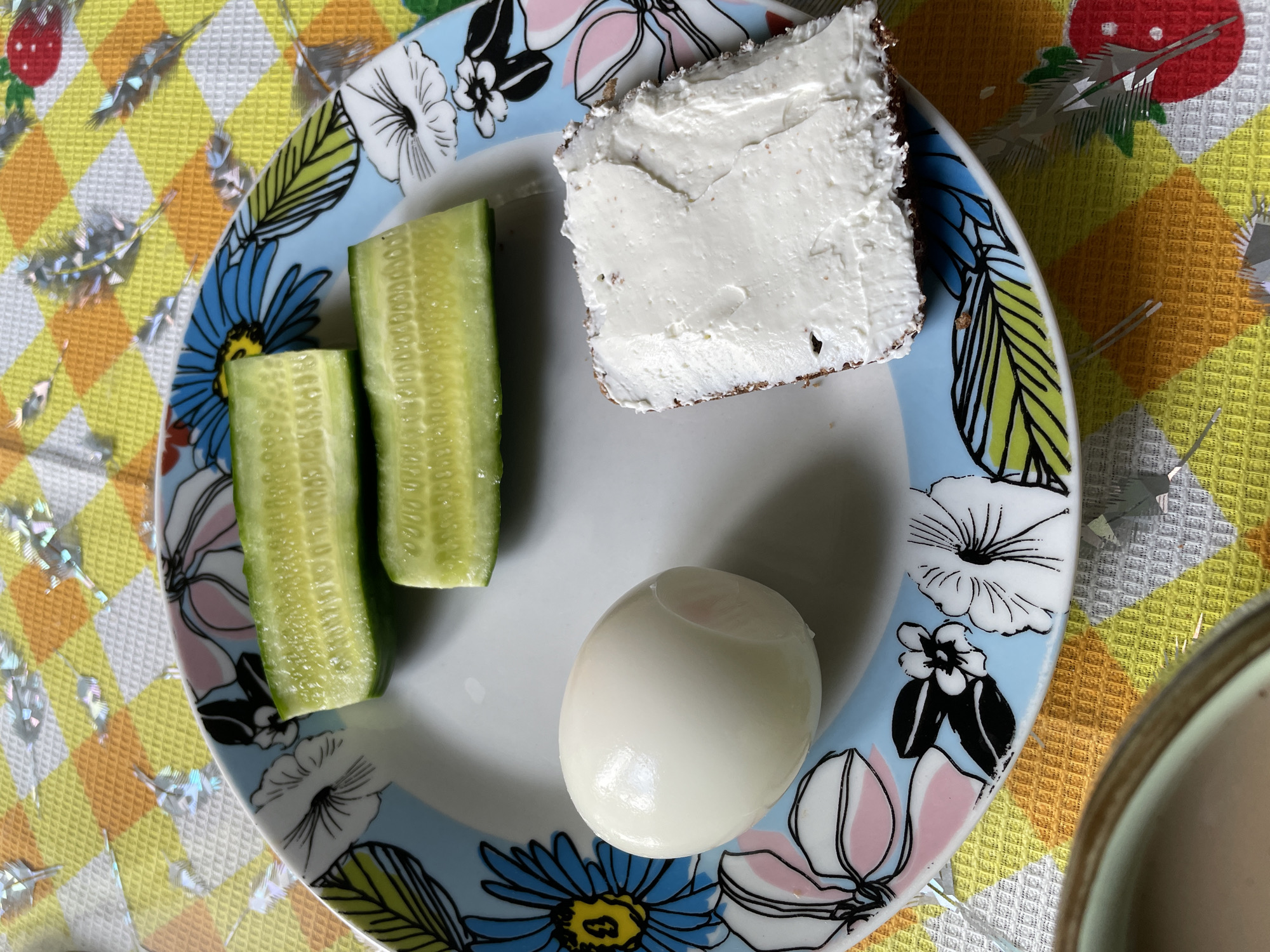 A floral-patterned plate rests on a yellow and white checkered surface, likely a tablecloth. On the plate are two cucumber segments, a peeled hard-boiled egg, and a square of bread thickly spread with white cream cheese. The setting appears to be indoors, potentially a kitchen or dining area. This image was taken in Talmenka, Russia. No people are visible in the image.