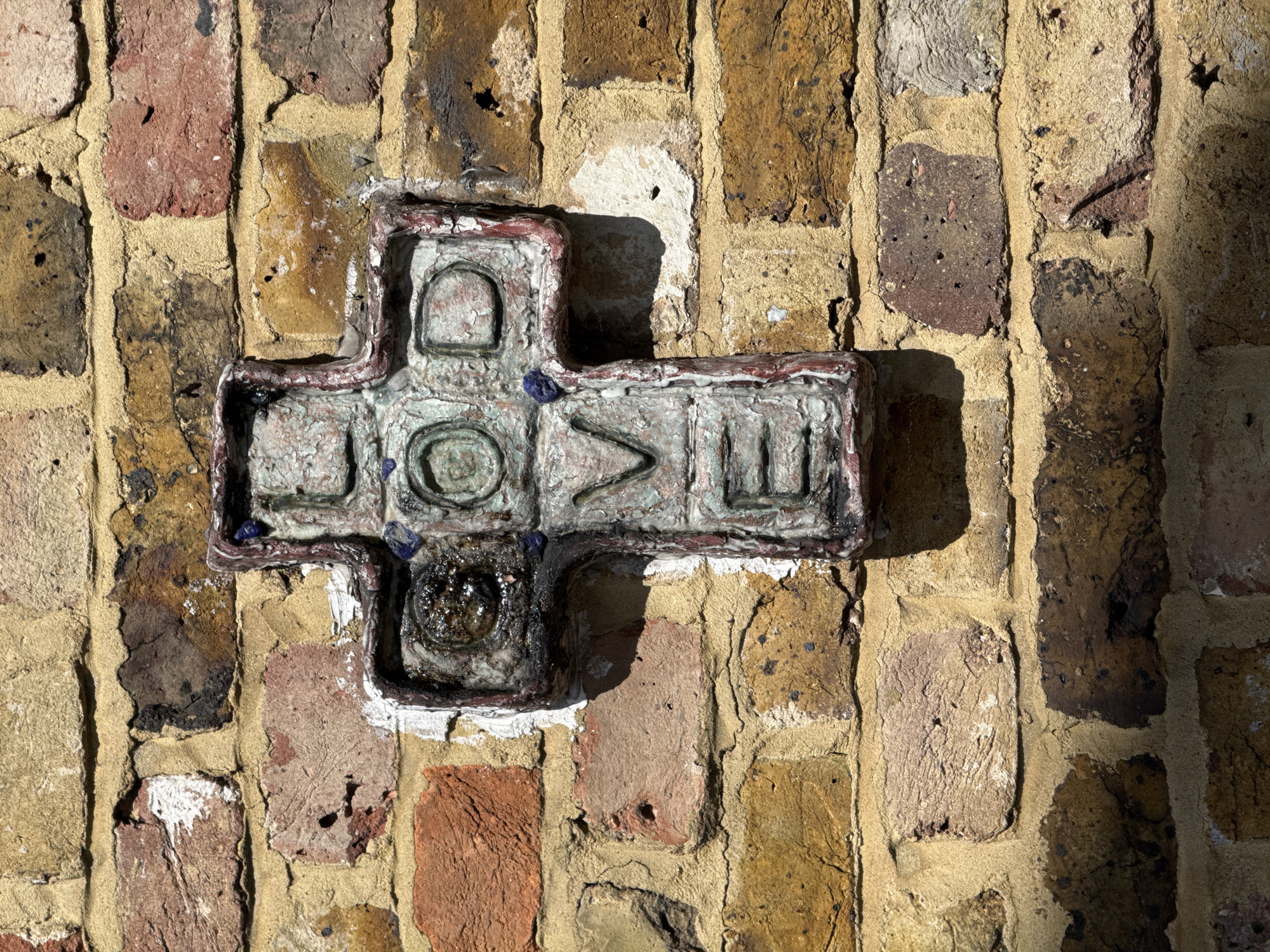 A cruciform plaque bearing the word "LOVE" is affixed to a brick wall. The plaque is made of a textured, possibly ceramic or plaster-like material, with a dark, weathered border and a pale interior with blue accents. The word "LOVE" is spelled out across the horizontal bar of the cross, with each letter occupying a separate section. The top and bottom sections of the cross are decorated with raised circular designs, the bottom one filled with dark resin. The brick wall has a variety of brick colors, including red, orange, and tan, and the mortar joints between the bricks are thick and textured. The scene is lit by sunlight, casting a shadow to the right of the plaque.
The image depicts a cross-shaped artifact that is actually a "love plaque" on a wall in London, United Kingdom. The work is by British artist Dscreet.