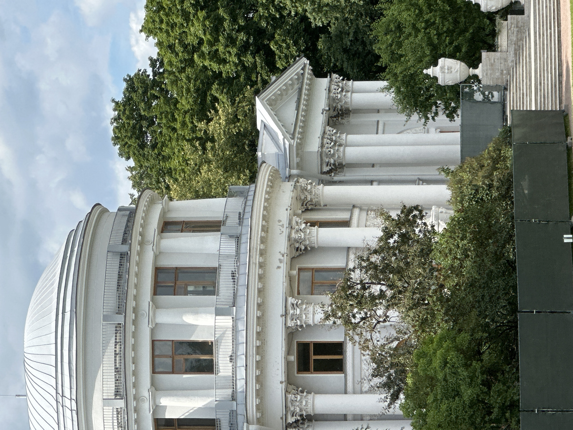 The image shows a portion of a large, white, classical-style building with a domed roof and numerous columns. The building is surrounded by lush green trees. In the foreground, there is a dark green fence and a set of stairs leading up to the building. A large decorative urn sits atop a stone pedestal on the right side of the stairs. The sky is partly cloudy. This building is located in Saint Petersburg, Russia, and appears to be a museum or a historical landmark.