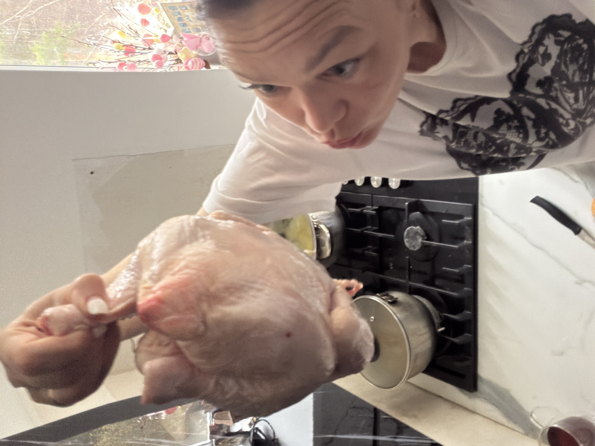 A person is holding a raw chicken in a kitchen. The person has short hair and is wearing a white t-shirt with a dark, possibly floral, design. The background includes a gas stovetop with a pot on it, and a white marble countertop. A window is visible in the background with branches and decorations. The exact location in the city of Chaikovsky, Russia, cannot be determined from the image.