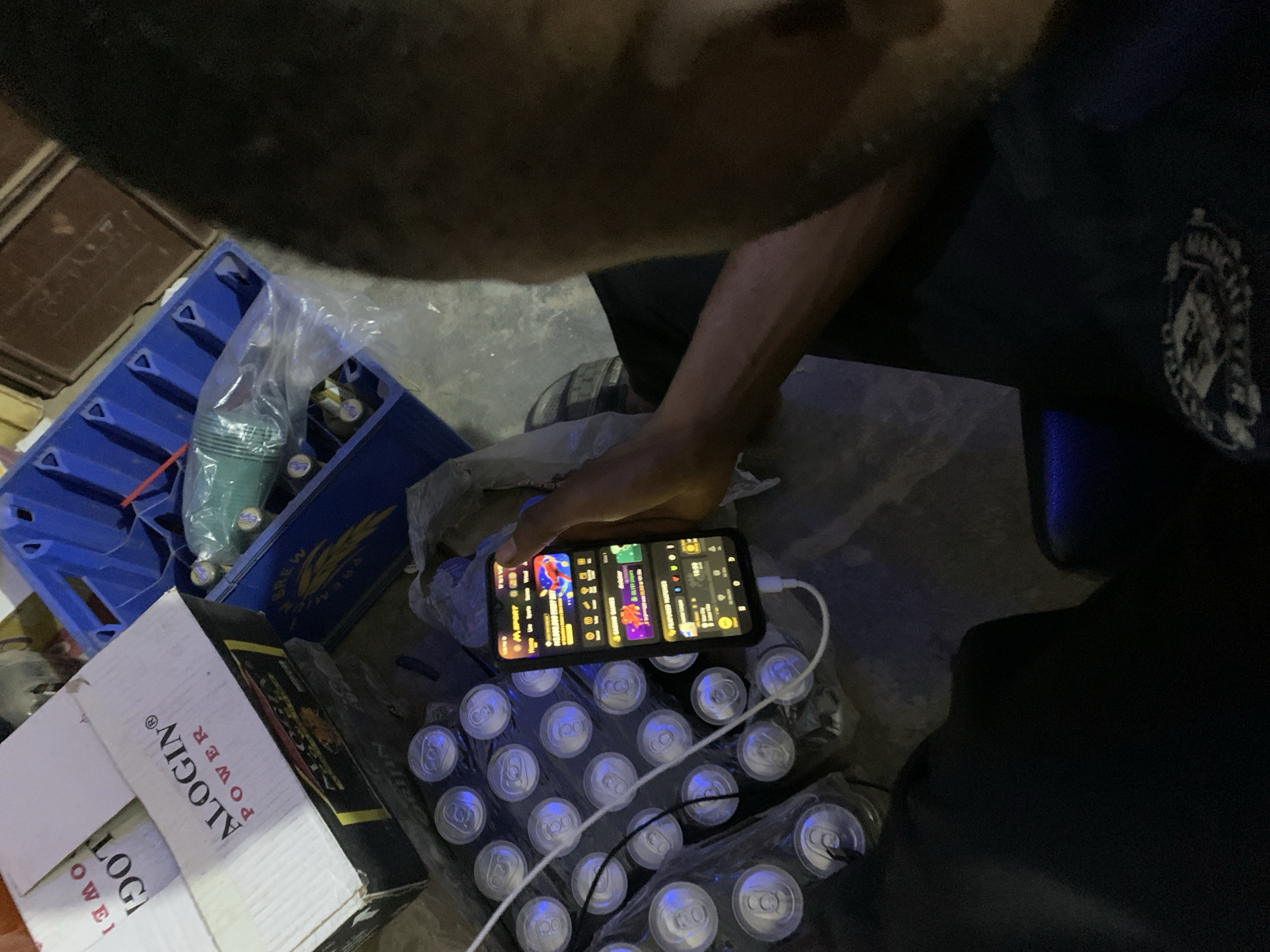 An overhead shot depicts a person interacting with a smartphone over a case of sealed beverage cans. The phone is charging. There is a blue beverage crate, a box labeled "ALOGIN POWER", and what appears to be a beverage in a plastic bag. The exact location within the city of New Stadium, Nigeria, is not discernible.
