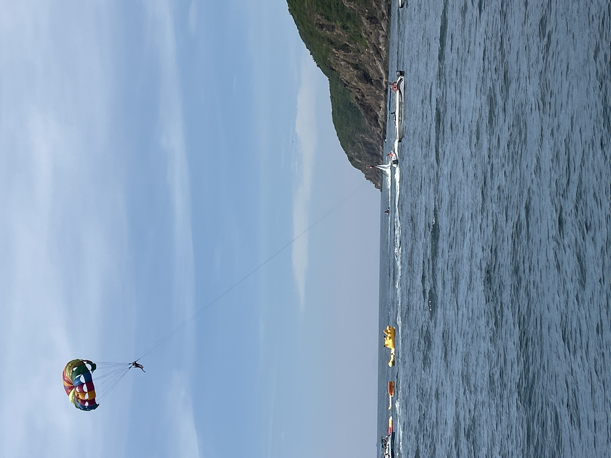A person is parasailing high above the ocean, attached to a colorful parachute. Below, several boats and watercraft are visible. To the left, a white boat with several passengers is moving. Further to the left and closer to the shore, a yellow inflatable tube is floating in the water, and another orange inflatable object is nearby. In the middle ground, a white speedboat is carrying passengers. To the right, a lush green, rocky island rises from the sea. The sky is blue with scattered clouds, suggesting a clear, sunny day. This scene appears to be in the waters near Nha Trang, Vietnam, known for its beaches and water sports.