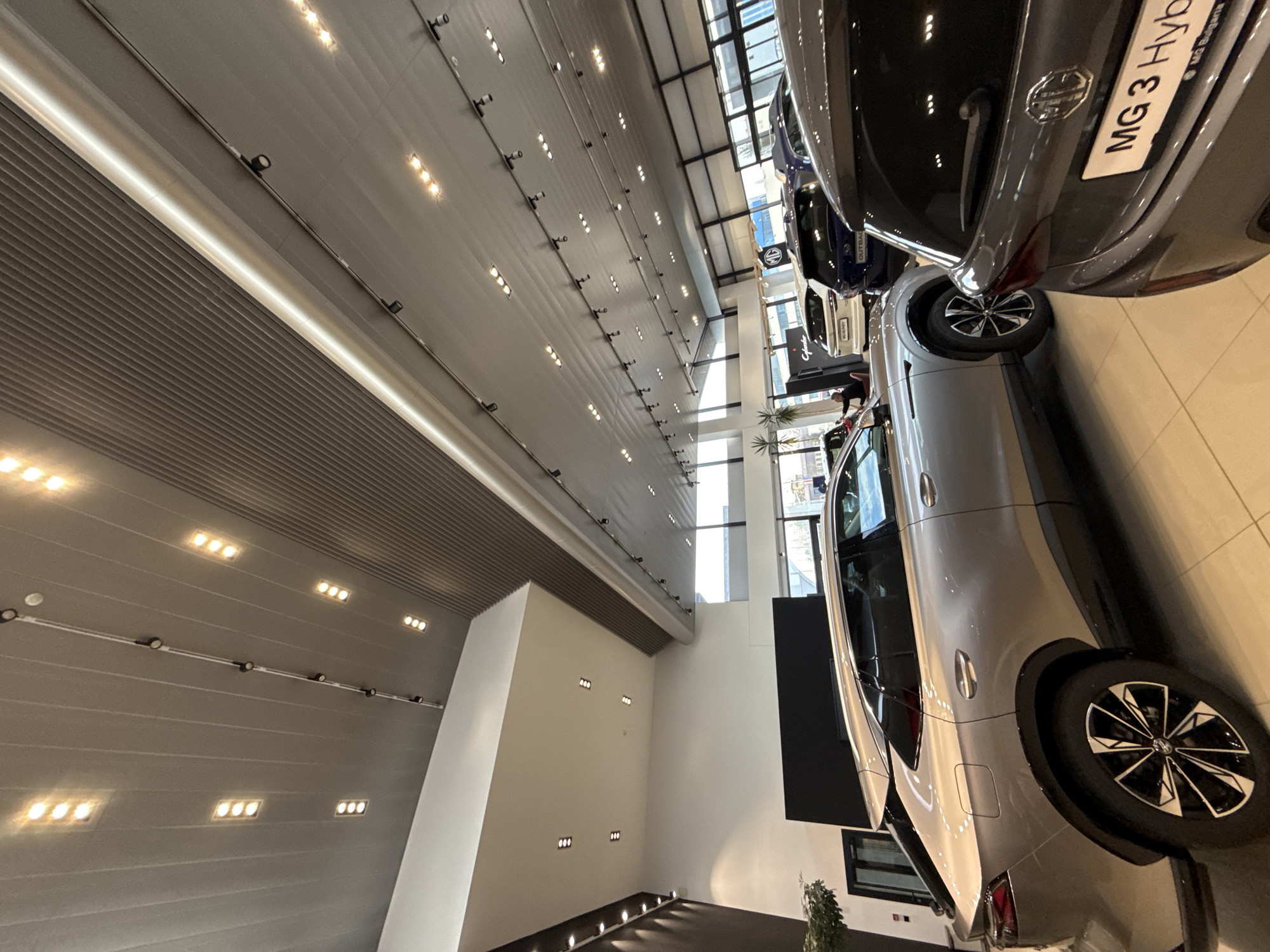 The image depicts the interior of the MG Motor Bulgaria car showroom located at бул. "Черни връх" 64, 1407 Hladilnika, Sofia, Bulgaria. The scene is a well-lit, modern automotive retail space with a high ceiling featuring a combination of horizontal ribbed grey panels, smooth white sections, and extensive lighting, including numerous recessed rectangular LED fixtures and a track lighting system with multiple spotlights. The floor is a light-toned, possibly cream-colored, tile surface.
In the foreground, the rear section of a dark grey MG 3 Hybrid hatchback is prominently displayed, showing its taillight and a Bulgarian license plate bearing the text "MG 3 Hybrid" and "MG Bulgaria". To its left, a light silver metallic coupe-SUV (possibly an MG ZS Coupe or MG One) is parked, showcasing its left rear quarter panel, rear wheel with multi-spoke alloy, and part of its passenger side. Behind this silver vehicle, a dark blue SUV, identified by faint text as a Subaru Outback, is visible, followed by a white SUV with a partially visible sign indicating "MG HS PHEV" in the background. An "MG" logo is visible on a pillar in the mid-ground.
One individual, dressed in dark attire, is partially visible in the mid-ground left, standing near the white SUV, appearing to observe the vehicles or an unseen display. Large floor-to-ceiling windows line the far wall of the showroom, offering a view of an exterior urban environment with distant buildings under natural light. A dark accent wall and a potted plant are visible on the far left.