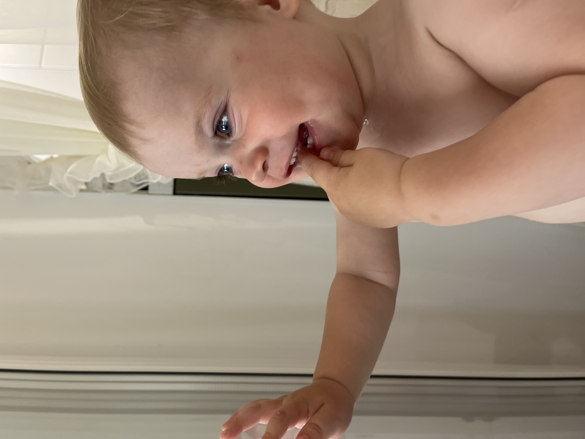 A young child is positioned close to the camera. The child is fair-skinned with blonde hair and blue eyes. The child is using their fingers to touch the front of their teeth. The background includes a shower curtain and what appears to be a window frame. The location is not apparent from this image.