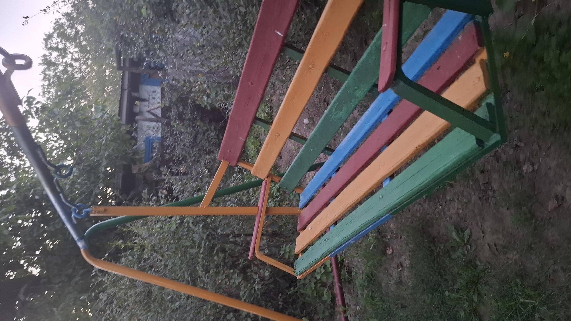 The image shows a vibrantly colored swing set with a bench seat in the foreground, situated outdoors. The swing set frame is primarily orange and green, with a dark gray horizontal bar at the top. The bench is constructed from wooden slats painted in alternating colors of green, blue, red, and orange. The background consists of dense foliage and trees, with a glimpse of a rustic structure, possibly a shed or small building, partially visible through the leaves. The ground appears to be a mix of dirt and sparse grass. The lighting suggests it is either early morning or late afternoon. The location is identified as Шұбарсу, Kazakhstan. There are no people visible in the image.