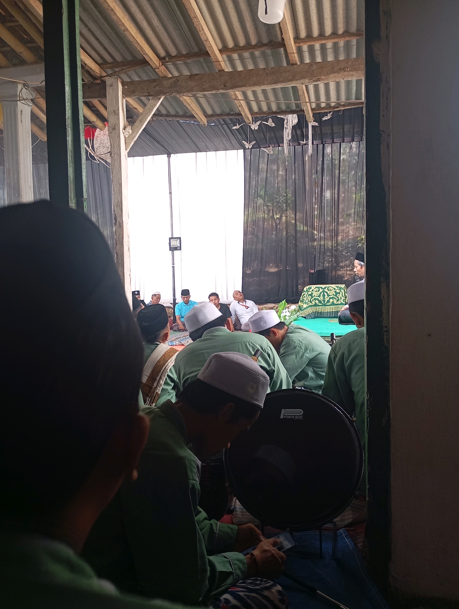 The image depicts an indoor gathering, likely a religious event, in Sumberjati, Indonesia. A group of men, some wearing peci (traditional Islamic caps) and green attire, are seated on the floor. In the foreground, the back of a person is visible, facing away from the camera, with a dark head covering. To the right, a large bass drum with the "POWER BEAT" logo is positioned. Further into the scene, several men are seated, some facing a central area where a man in a white shirt and a blue shirt are present. A raised platform draped with green fabric and adorned with an ornate pattern is visible in the background, behind which a black curtain hangs, revealing a silhouette of trees. The structure appears to be a simple, open-air building with a corrugated metal roof supported by wooden beams. The overall atmosphere suggests a communal gathering, possibly a prayer service or religious lecture.